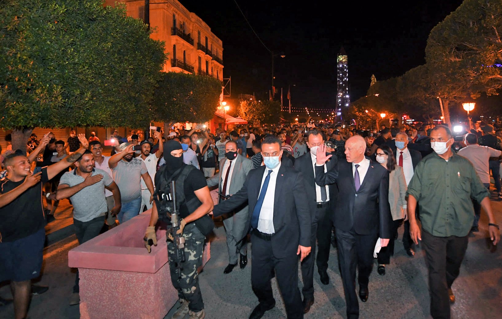 این تصویر گروهی از مردم را در یک خیابان در شب نشان میدهد که در حال راهپیمایی هستند. در میان آنها، یک فرد با لباس رسمی و گروهی از افراد دیگر حضور دارند. همچنین، یکی از افراد مسلح با یونیفورم مشخص در آنجا دیده میشود. به نظر میرسد که این صحنه به یک تجمع یا تظاهرات مرتبط باشد و تعدادی از معترضان و شخصیتهای دیگر در حال تعامل هستند. نورهایی از چراغهای خیابانی به وضوح در صحنه قابل مشاهده است.