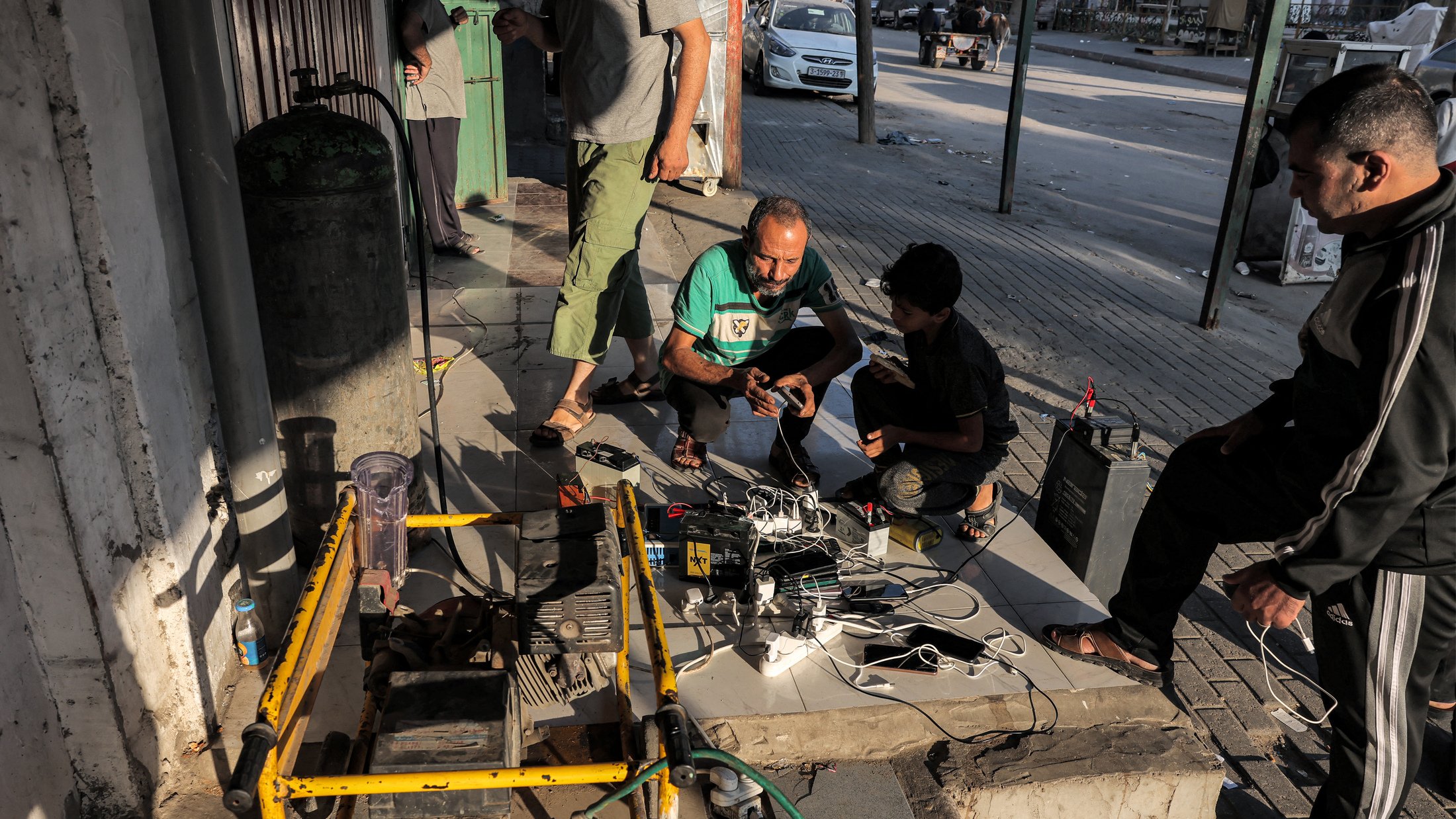 در تصویر، گروهی از افراد در حال کار بر روی یک دستگاه یا تجهیزات الکتریکی در خارج از یک ساختمان هستند. تعدادی سیم و کابل در اطراف آنها وجود دارد و به نظر میرسد که مشغول تعمیر یا بررسی یک دستگاه هستند. در پسزمینه، خیابانی دیده میشود و تعدادی وسیله نقلیه نیز در حال عبور هستند. جو تصویر به صورت غیررسمی و مشغول به کار به نظر میرسد.