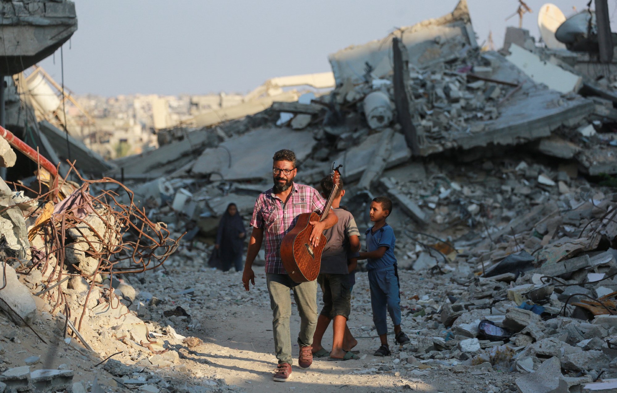 تصویر یک مرد را نمایش میدهد که در حال حرکت در میان ویرانههای یک منطقه تخریبشده است. او یک گیتار به دوش دارد و به نظر میرسد در حال نواختن یا آماده نواختن موسیقی باشد. دو کودک نیز به او نزدیک هستند، یکی از آنها به مرد وابسته است و به نظر میرسد او را دنبال میکند. در پسزمینه، آوار و خرابههای ساختمانها دیده میشود که نشانهای از یک بحران یا جنگ را نشان میدهد. نورخورشید در آسمان دیده میشود که ممکن است تصاویری از امید را القا کند.