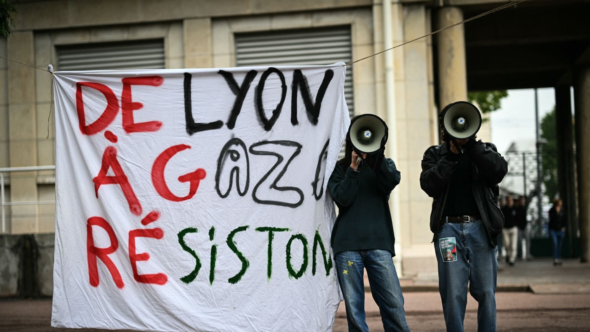 تصویر نشاندهنده دو فرد است که در کنار یک بنر بزرگ ایستادهاند. بنر با رنگهای مشکی، قرمز و سبز نوشته شده و متن آن به زبان فرانسوی است. همچنین، هر یک از این افراد یک بلندگو در دست دارند و به نظر میرسد در حال اعتراض یا ابراز نظر هستند. پسزمینه تصویر به رنگهای خنثی و ساختمانی است که نشاندهنده یک فضای شهری میباشد.