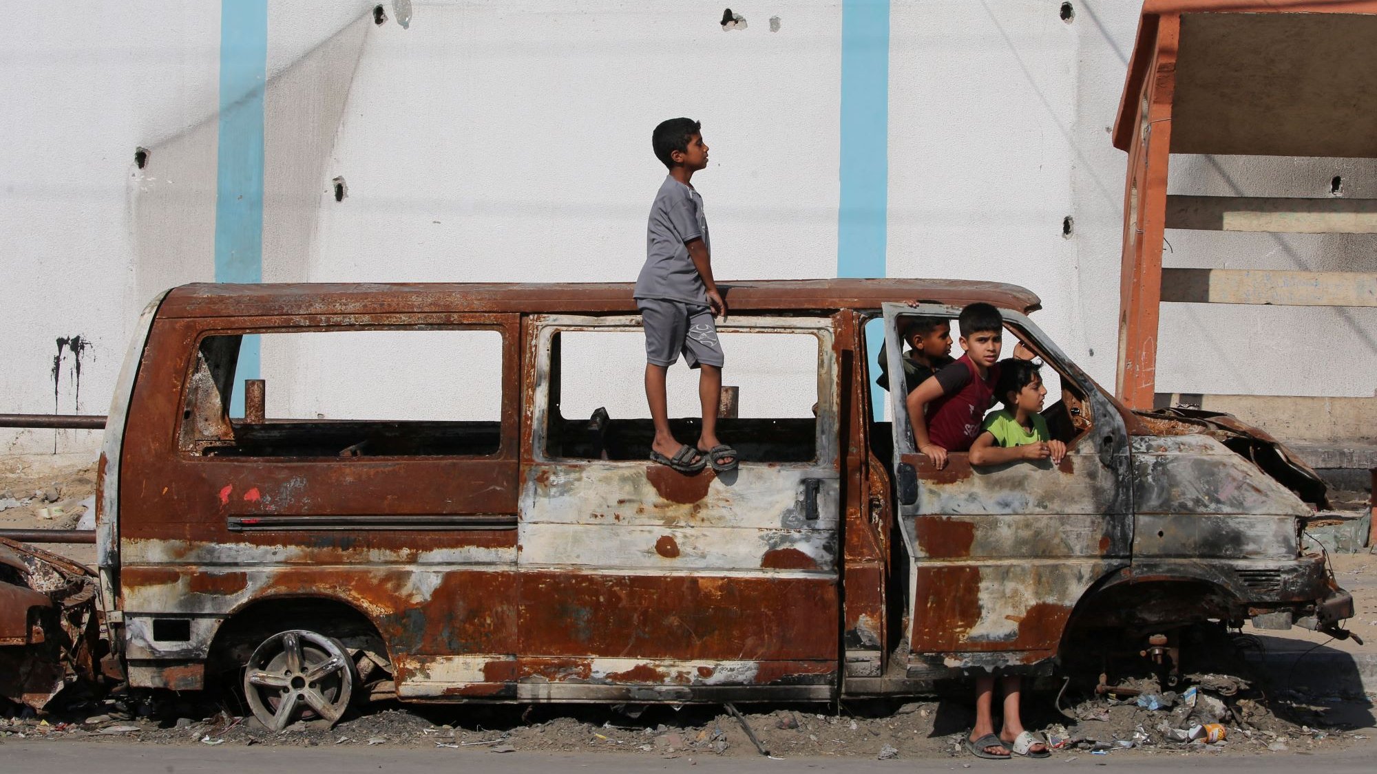 در این تصویر، یک خودرو قدیمی و زنگزده دیده میشود که در یک محیط شهری و آشفته قرار دارد. چند کودک در اطراف این خودرو بازی میکنند و یک کودک بر روی سقف آن ایستاده است. مناظر پشت سر آنها شامل دیوارهای آسیبدیده و ناپایداریهای دیگر است که نشاندهنده شرایط سخت و فرسوده منطقه است. این تصویر احساساتی از کودکی و بازی در شرایط نامناسب را منتقل میکند.