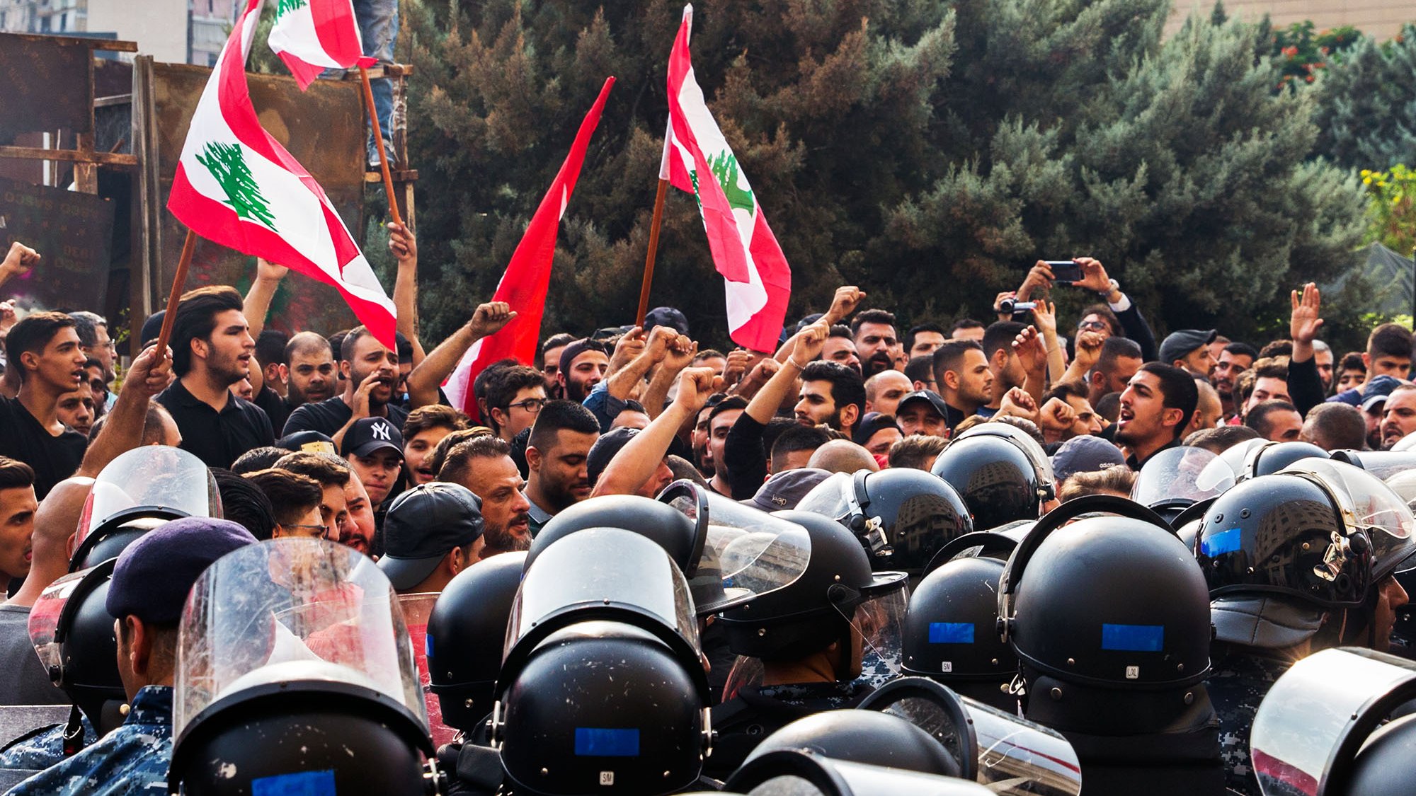در این تصویر، یک تجمع اعتراضی در لبنان دیده میشود. گروهی از مردم در حال شعار دادن و بهسوی دوربین نگاه میکنند، در حالی که پرچمهای لبنان را در دست دارند. در مقابل آنها، گروهی از نیروهای پلیس با لباسهای ضدشورشی و کلاهخودهایی مشغول محافظت از جایگاه هستند. فضای تصویر تنشآلود و احساسات قوی در بین معترضین و نیروهای پلیس به وضوح حس میشود. در پسزمینه، ساختمانها و درختان وجود دارند که به جو عمومی صحنه اشاره میکنند.