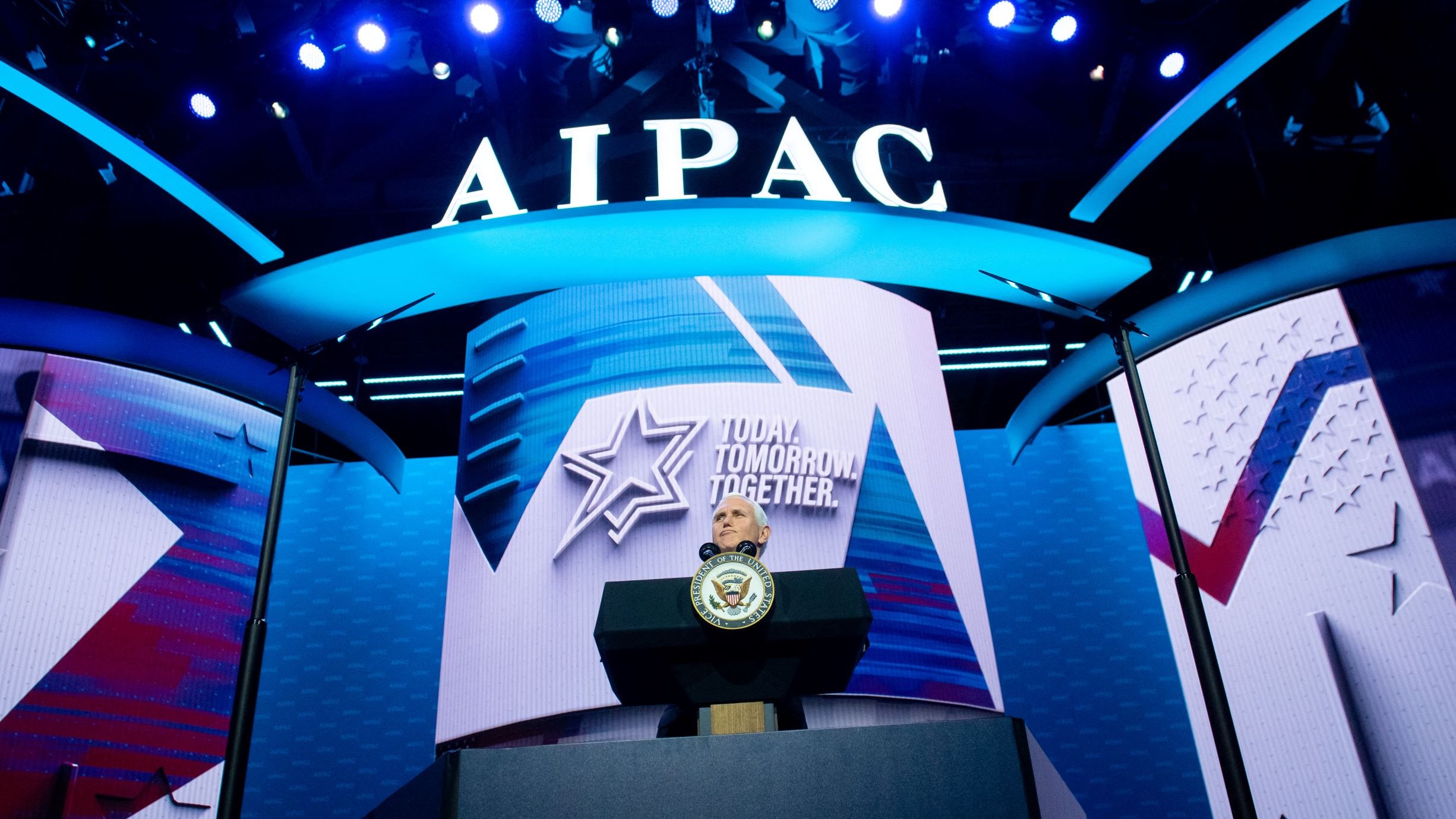 تصویر یک صحنه از یک کنفرانس یا گردهمایی است. در پسزمینه، یک نمایشگر بزرگ با نام "AIPAC" و شعار "Together Tomorrow" دیده میشود. در جلوی صحنه، یک میز سخنرانی وجود دارد که بر روی آن نشان رسمی (احتمالاً نشان کشور یا دولت) قرار دارد. نورپردازی آبی و سفید به فضایی بانشاط و رسمی بخشیده است. طراحی کلی صحنه نشان دهنده یک رویداد مهم است.