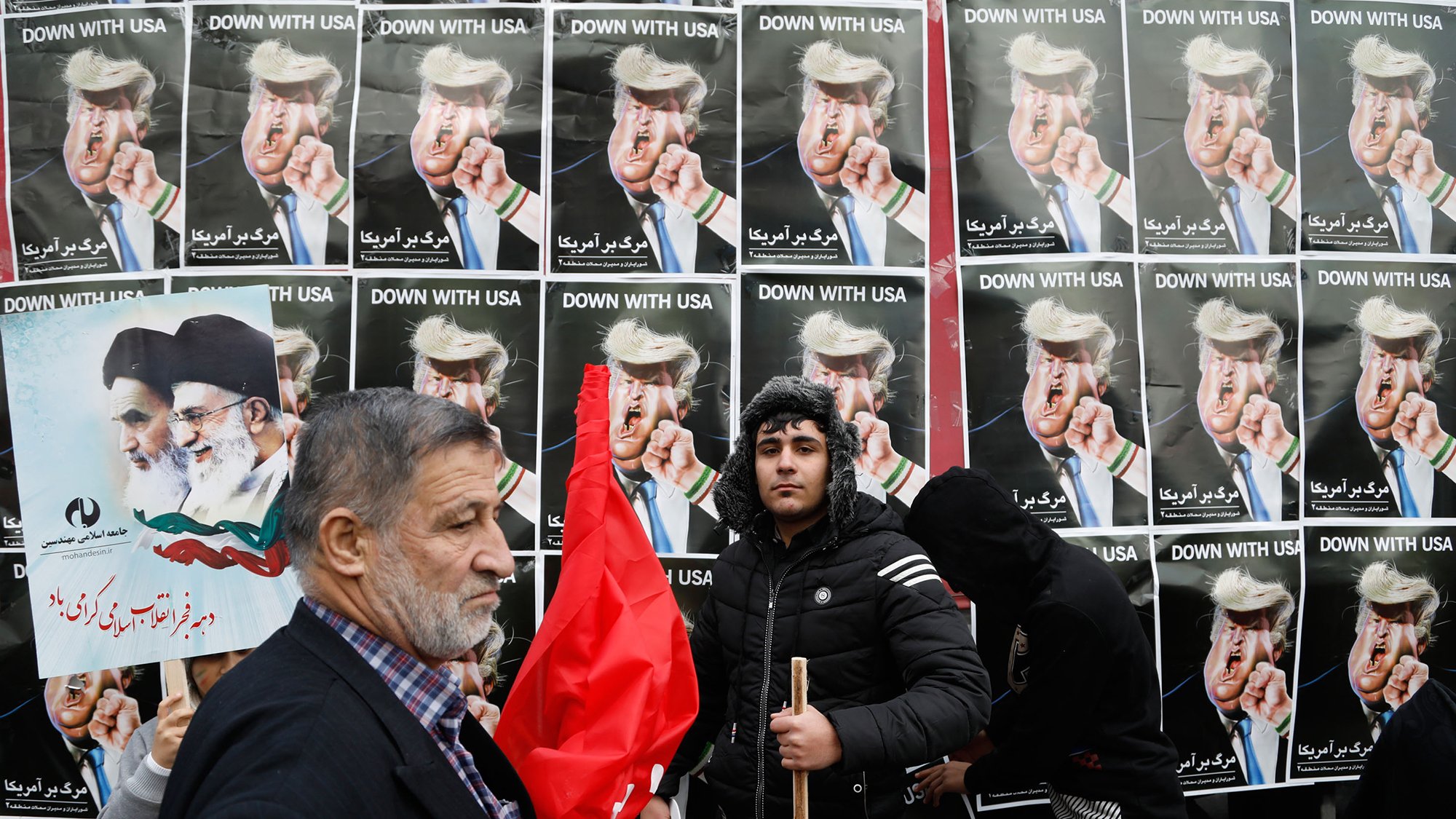 در این تصویر، گروهی از مردم در حال تظاهرات به نمایش گذاشتهاند. در پسزمینه، پوسترهایی با تصاویر رهبران سیاسی و متنی به زبان فارسی دیده میشود که به موضوعی خاص اشاره دارد. یکی از افراد به سمت دوربین نگاه میکند و فرد دیگری با یک چوب در دست و لباسی سیاه در کنار او ایستاده است. برخی از افراد دیگر نیز در این صحنه حضور دارند که نشاندهنده جو تظاهرات است. پرچمها و پلاکاردها نیز در میان جمعیت دیده میشود که نشاندهنده احساسات و پیامهای سیاسی آنها است.