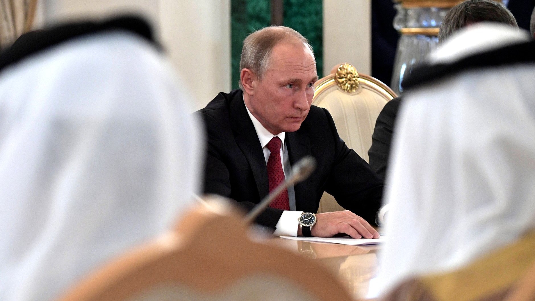 در این تصویر، فردی که به نظر میرسد ولادیمیر پوتین است، در حال نشستن در یک جلسه به سمت جلو نگاه میکند. او کت و شلوار مشکی به همراه یک رابعه قرمز به تن دارد. در اطراف او، چهرههایی از دیگر شرکتکنندگان در جلسه دیده میشود، اما جزئیات آنها قابل تشخیص نیست. فضای عمومی جلسه جدی به نظر میرسد و میز مستطیلی به همراه دکوراتورهای رسمی دیده میشود.