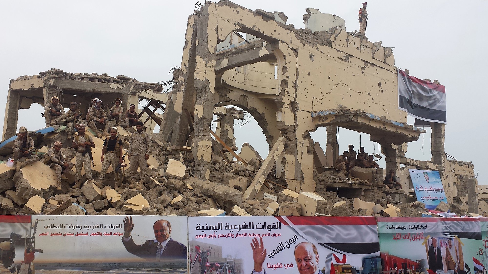 این تصویر نمایی از یک ساختمان ویرانشده را نشان میدهد که تخریب آن به وضوح قابل مشاهده است. در پسزمینه، تعدادی نظامی به چشم میخورند و روی دیوارهای ویرانه بنرهایی وجود دارد که به نظر میرسد تبلیغاتی سیاسی هستند. در این بنرها، تصاویری از پروژهها و شخصیتهای سیاسی دیده میشود. به طور کلی، تصویر به احساساتی از جنگ، تخریب و همچنین تبلیغات سیاسی اشاره میکند.