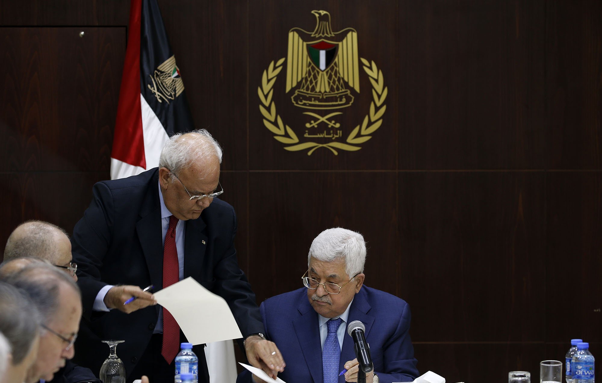 در این تصویر، یک جلسه رسمی در حال برگزاری است. در مرکز تصویر، مردی به نام محمود عباس نشسته است که به نظر میرسد در حال مطالعه مدارک یا مستندات است. افراد دیگری نیز در اطراف او نشستهاند. یکی از آنها به سمت عباس خم شده و به او مدرکی نشان میدهد. در پسزمینه، پرچم فلسطین و نماد رسمی مرتبط با دولت فلسطین دیده میشود، که به فضایی رسمی و سیاسی اهمیت میدهد. میز گردی که آنها دور آن نشستهاند، با لوازم اداری و بطریهای آب تزیین شده است.