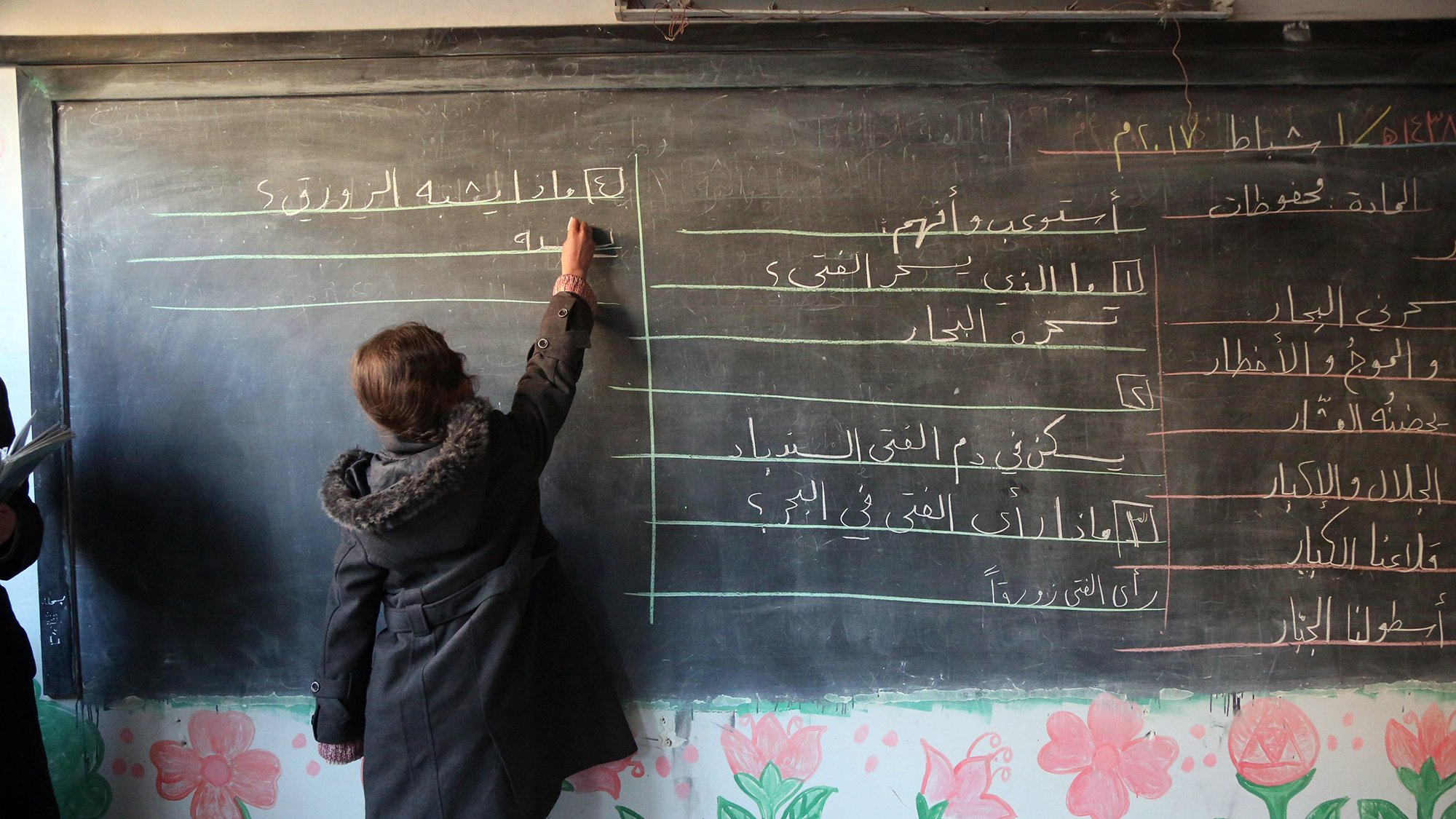 در این تصویر، یک دانشآموز در حال نوشتن روی تخته سیاه در کلاس درس است. او با دقت در حال نوشتن مطالبی است که به نظر میرسد مربوط به درس خاصی باشد. در پسزمینه، دیوار کلاس با نقاشیهای رنگی و گلها تزئین شده است. این فضا نمایانگر یک محیط آموزشی است که در آن دانشآموزان مشغول یادگیری هستند.