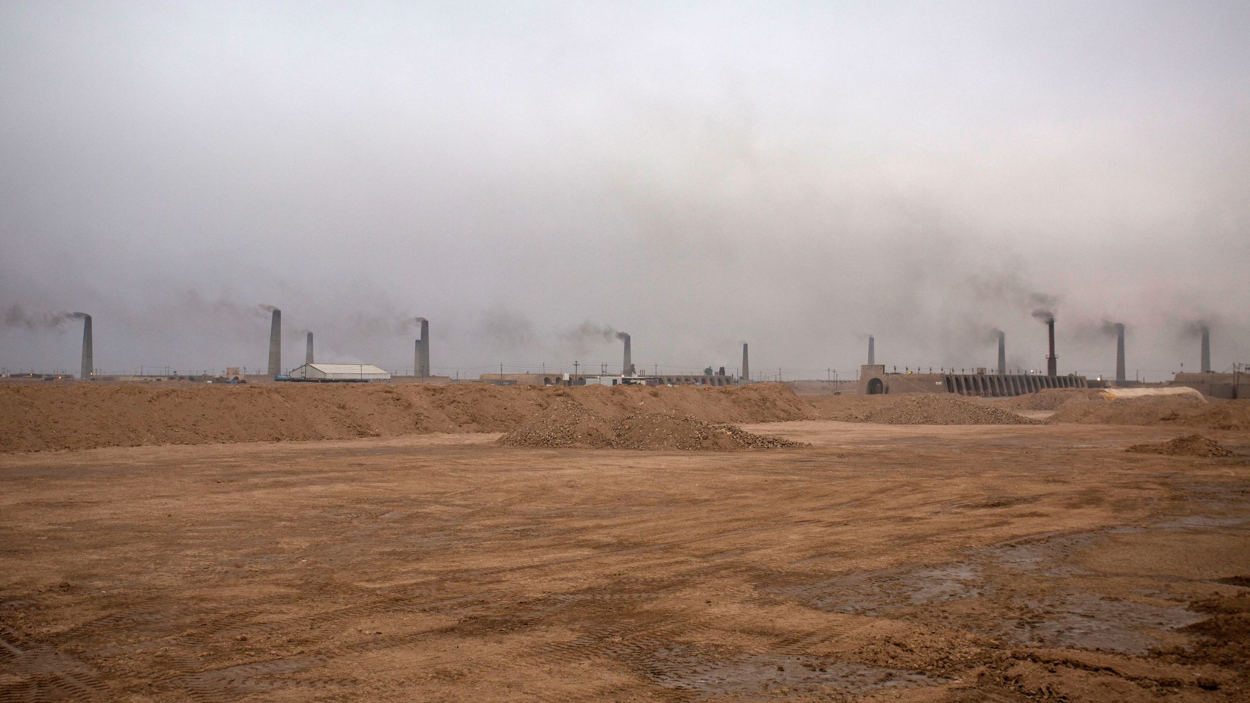 این تصویر یک محیط صنعتی را نشان میدهد که در آن دود از دودکشهای بلند بیرون میآید. آسمان به طور کلی ابری و خاکستری به نظر میرسد و هوای منطقه آلوده است. زمین خاکی و خشک به نظر میرسد و هیچ گونه پوشش گیاهی قابل مشاهده نیست. این صحنه میتواند نشانگر فعالیتهای صنعتی و مشکلات زیستمحیطی باشد.