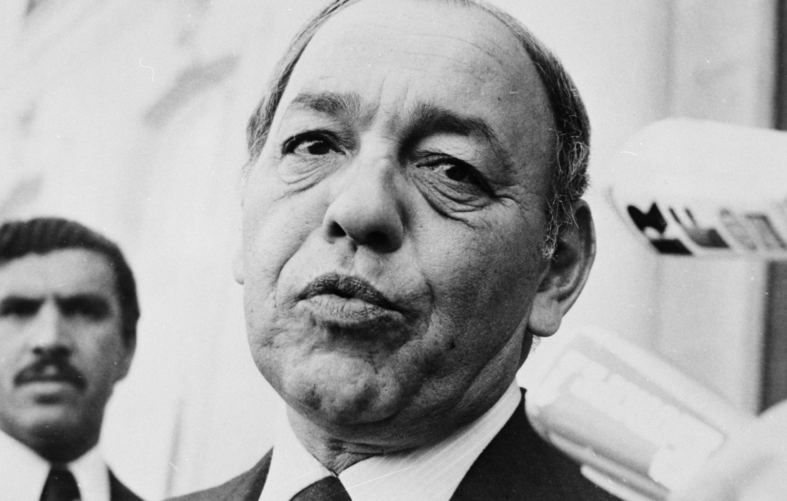 این تصویر یک مرد میانسال را نشان میدهد که ظاهری جدی و متمرکز دارد. او لباس رسمی به تن دارد و به دوربین نگاه میکند. در پسزمینه، شخص دیگری نیز دیده میشود که به نظر میرسد در حال نظارت یا همراهی با او است. فضای تصویر به طور کلی رسمی و مرتبط با یک موقعیت اجتماعی یا سیاسی به نظر میرسد.