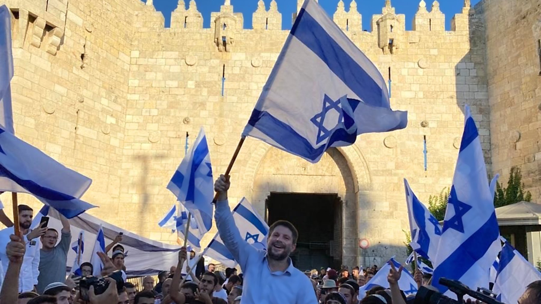 تصویر نشاندهنده جمعیتی است که با پرچمهای اسرائیل در حال جشن و شادی هستند. در میان این جمعیت، فردی در حال بلند کردن پرچم و ابراز شادی است. پسزمینه تصویر یک دروازه تاریخی و دیوار سنگی نمایان است که نشاندهنده یک مکان مهم است. افراد حاضر در تصویر لباسهای مختلفی به تن دارند و فضای کلی عکس نشاندهنده شور و نشاط است.
