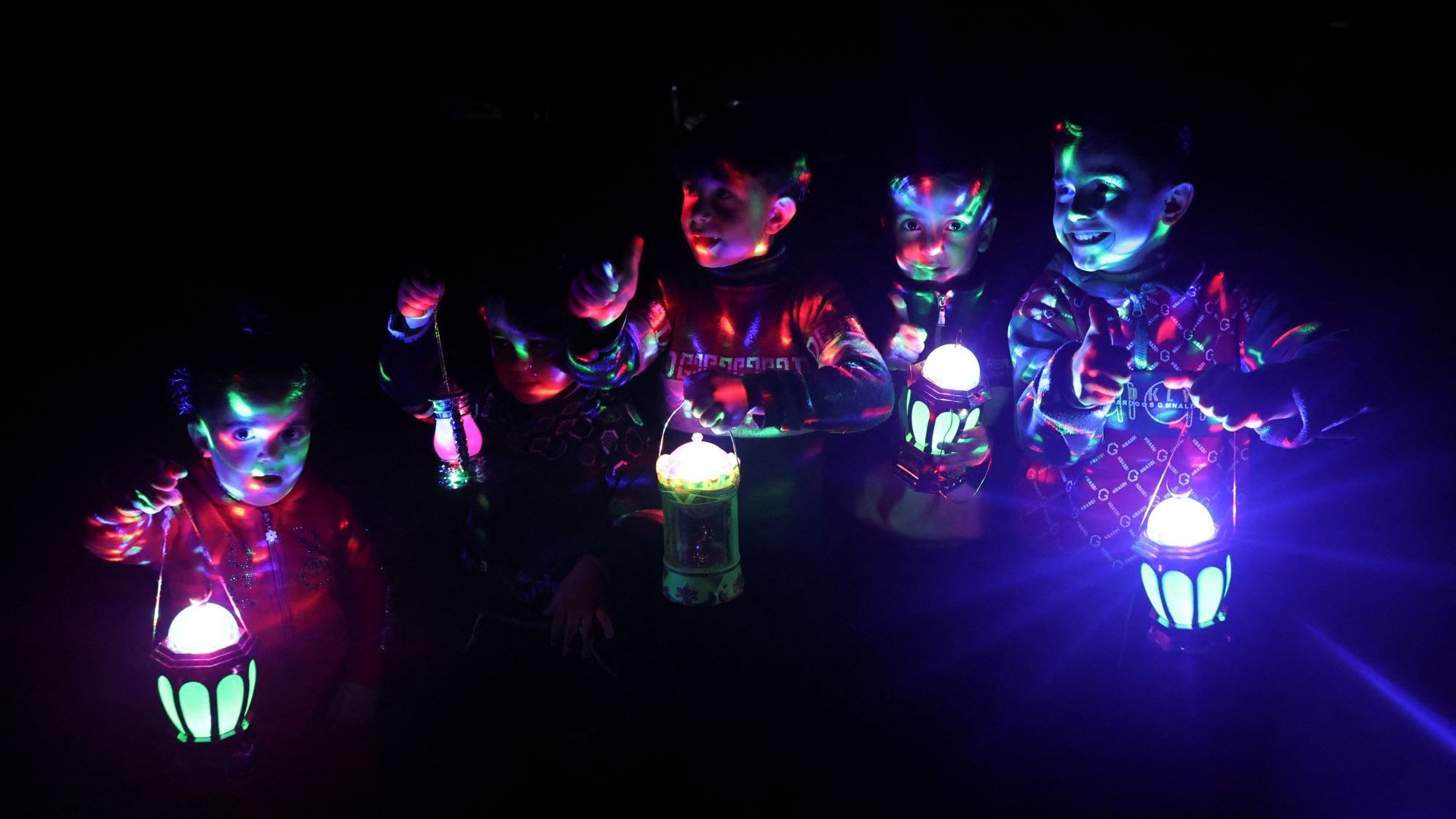 در این تصویر، چند کودک در یک محیط تاریک ایستادهاند و هرکدام در دست خود یک فانوس با نورهای رنگی دارند. نورهای فانوسها به زیبایی در فضای اطراف پخش شده و چهرهها و بدنهای کودکان را به طرز جالبی روشن کردهاند. کودکان در حال شادی و بازی هستند و به نظر میرسد که لحظاتی سرگرمکننده را سپری میکنند. رنگهای درخشان و جلوههای نوری به تصویر حالت جشن و شادی بخشیدهاند.