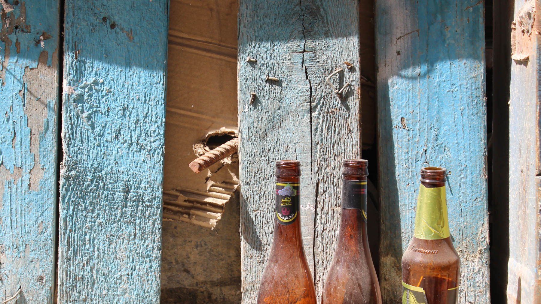 در این تصویر، سه بطری خالی در کنار دیواری که رنگ آبی دارد و کمی فرسوده به نظر میرسد، قرار گرفتهاند. دیواره دارای نواحی ترکخورده و برخی زواید و نقصهاست. بطریها به صورت نامرتب در کنار هم قرار دارند و بافتی کهنه و رنگهایی مات دارند، که به تصویر حس قدیمی و رها شدهای میبخشد. در پسزمینه، تکههایی از دیوار نیز قابل مشاهده است.