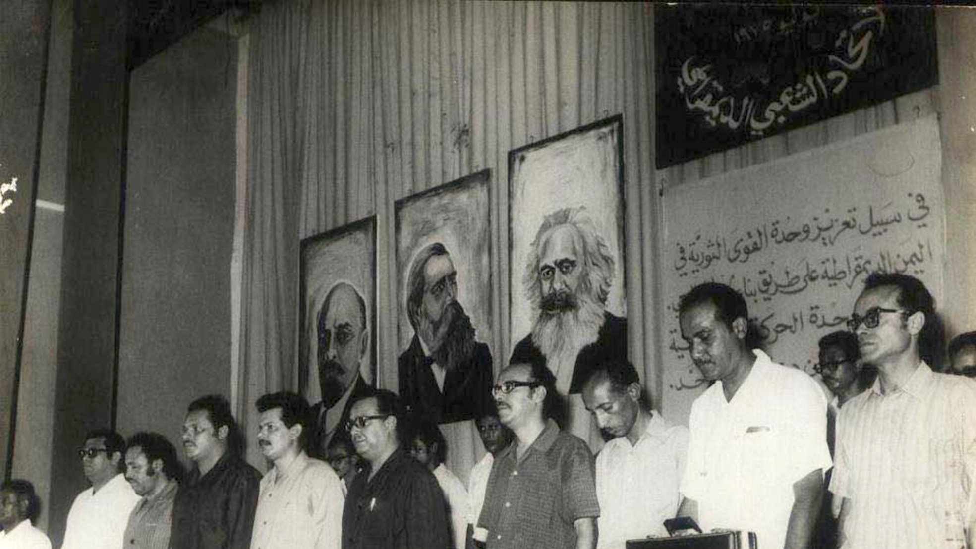 این تصویر مربوط به یک تجمع یا کنفرانس سیاسی به نظر میرسد. در پسزمینه، تابلوهایی از چهرههای معروف تاریخی یا سیاسی وجود دارد. افرادی که در جلوی صحنه ایستادهاند، موضع جدی و متفکرانهای دارند و در حال گوش دادن به سخنرانی یا برنامهای هستند. فضای کلی تصویر به نظر میرسد که تمرکز بر مسائل اجتماعی و سیاسی داشته باشد. دیوار پشتشان نیز با متنی به زبان عربی تزئین شده است.