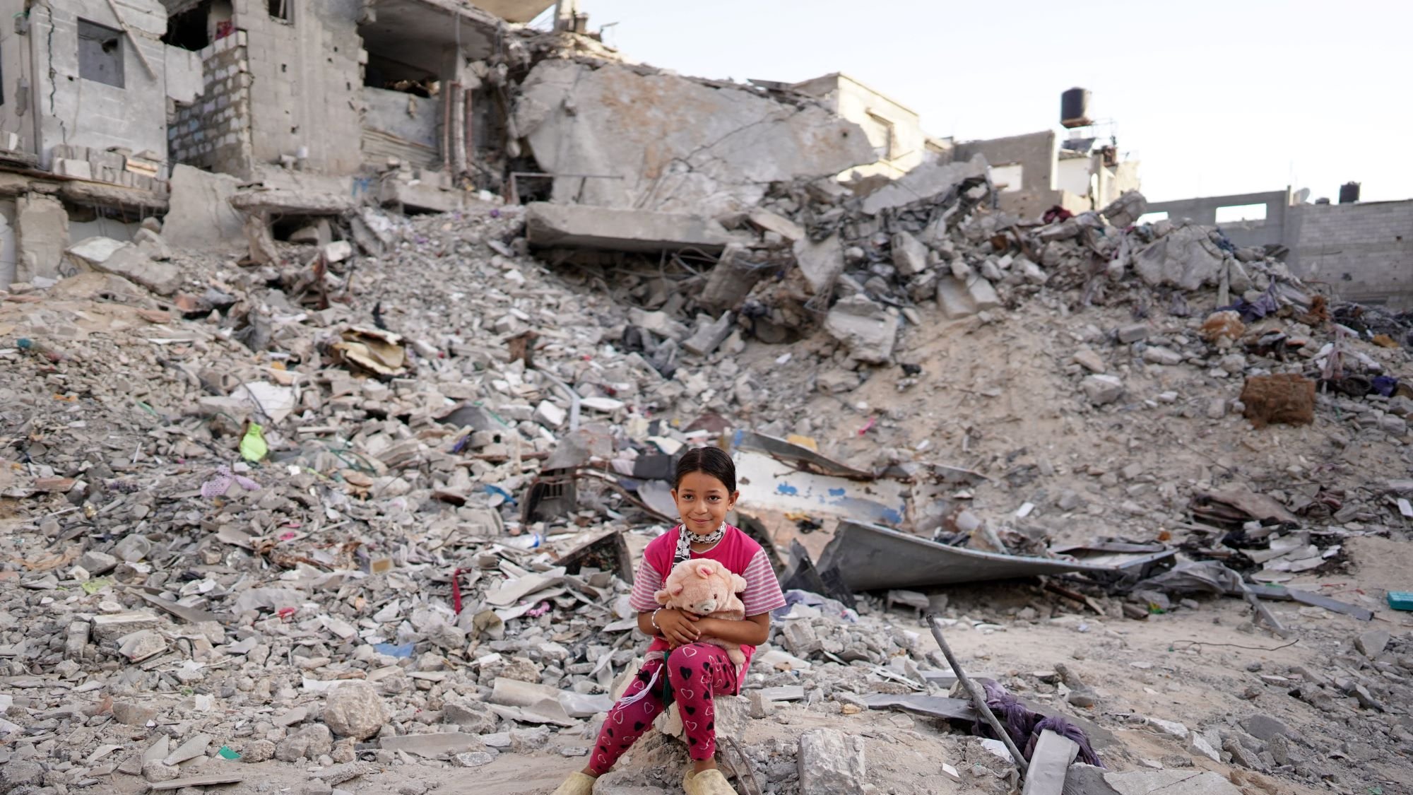 در این تصویر، دخترکی کوچک بر روی زمین در میان ویرانههای یک ساختمان نشسته است. در پسزمینه، تخریبهای وسیع و انبوهی از آجرها و مصالح ساختمانی دیده میشود. دخترک با لباسی صورتی و شلوار آبی، عروسکی در آغوش دارد و به دوربین نگاه میکند. وضعیت اطراف او به وضوح نشاندهنده ویرانی و داغی ناشی از یک حادثه یا جنگ است. احساساتی از معصومیت و ناامیدی در این تصویر قابل مشاهده است.