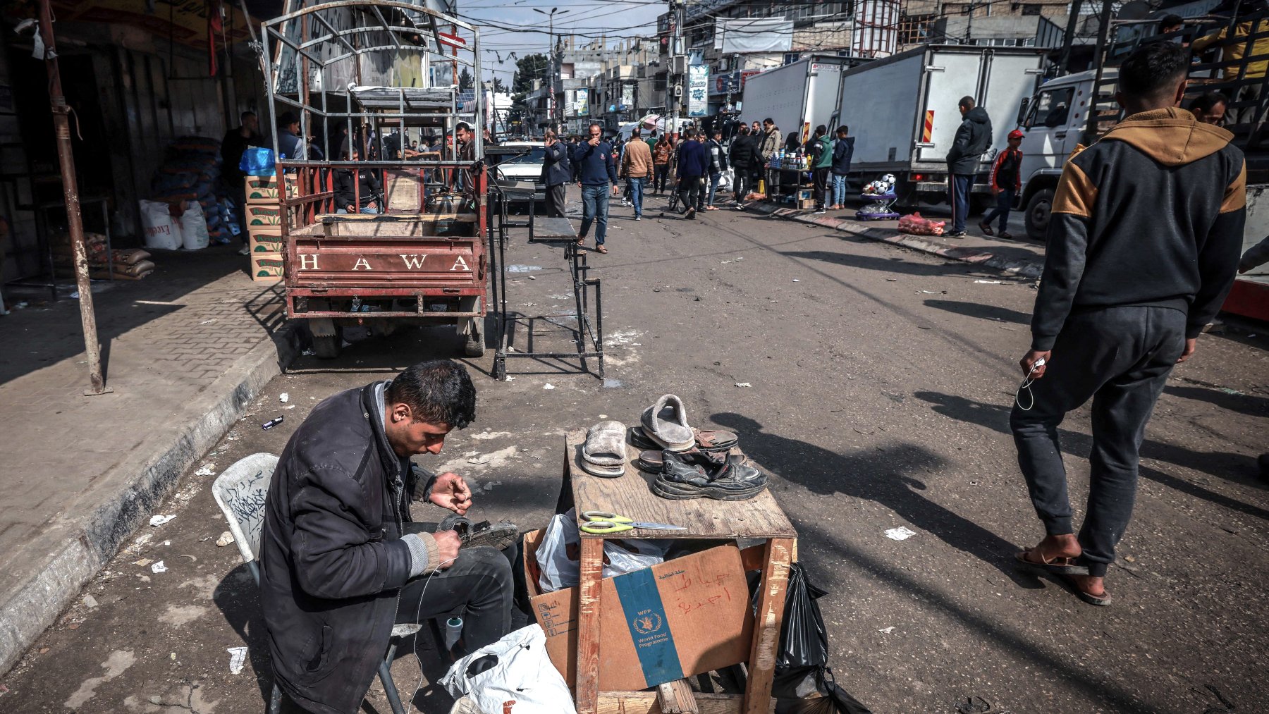 در تصویر، مردی در حال تعمیر یا واکس زدن کفشها است. او روی یک صندلی نشسته و وسایل کارش را بر روی یک میز کوچک چوبی قرار داده است. در اطراف او، افرادی در حال عبور و مرور هستند و تعدادی خودروی باربری در پسزمینه دیده میشود. فضایی شلوغ و شهری به نظر میرسد و فعالیتهای روزمره در حال انجام است. چند کشیده و بقیه اجناس نیز در اطراف قرار دارند.