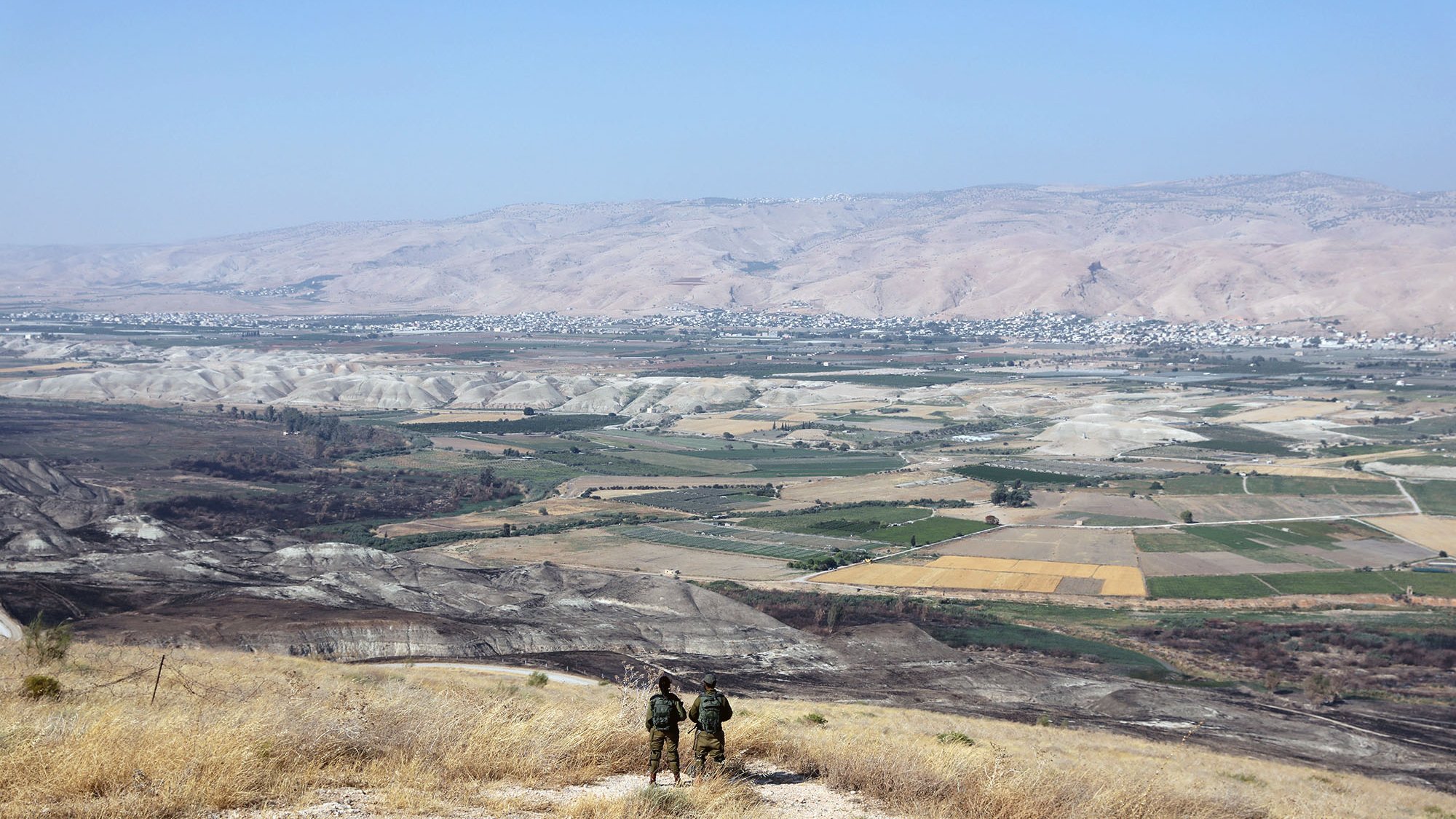 در این تصویر، دو نفر در حال ایستادن بر روی یک تپه یا ارتفاع هستند و به منظرۀ وسیع و زیبای زیر پای خود نگاه میکنند. در زیر، دشتها و زمینهای کشاورزی با رنگهای مختلف دیده میشوند. دامنههای کوهها در دوردست قرار دارند و آسمان آبی و صاف به چشم میخورد. فضا به نظر آرام و دلنشین میرسد و نشاندهندهٔ مناظر طبیعی و انسانی در منطقهای کوهستانی است.