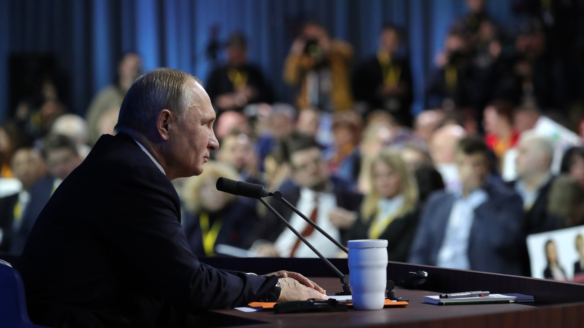 در این تصویر، ولادیمیر پوتین، رئیسجمهور روسیه، در حال سخنرانی یا پاسخ به سؤالات در یک کنفرانس مطبوعاتی به نظر میرسد. او در حال نشستن در کنار یک میز است که میکروفون دارد و در مقابل او تعدادی از خبرنگاران و حضار به نظر میرسند. محیط اطراف کمی تاریکتر است و تمرکز اصلی بر روی پوتین قرار دارد. تعدادی از افراد حاضر در جمع در پسزمینه دیده میشوند.
