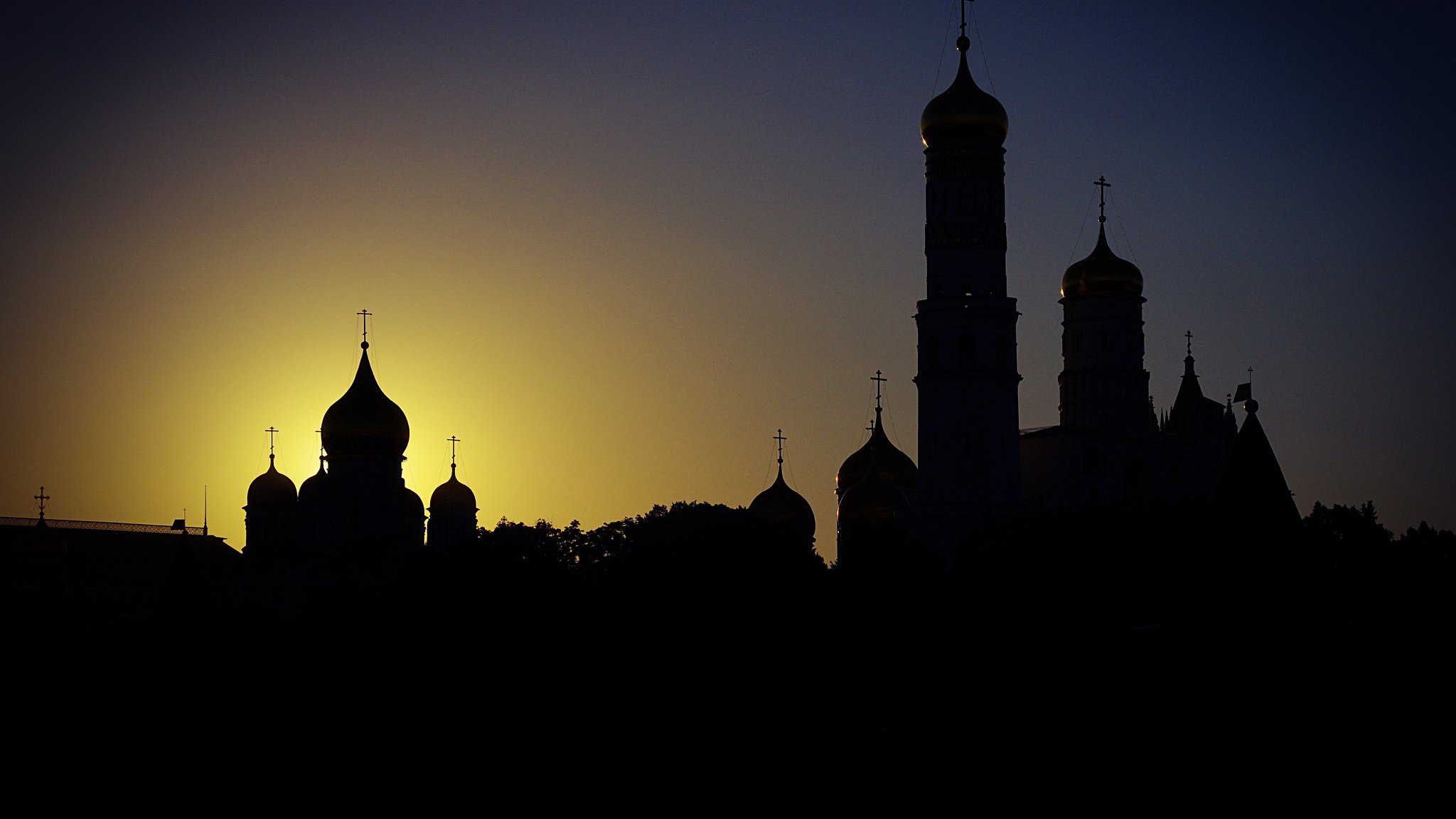 این تصویر نمایی از یک افق زیبا در هنگام غروب را نشان میدهد. در پسزمینه، خورشید به تدریج در حال غروب است و نور طلایی آن به صورت مجسمهای در آمده که بر روی قلههای ساختمانها تابیده میشود. در جلو، ساختمانهای مذهبی با گنبدها و برجهای بلند، به صورت سیاهچرمی در برابر نور زمینه قرار دارند. سایههای این بناها، محیط را به فضایی رازآلود و باشکوه تبدیل کردهاند.