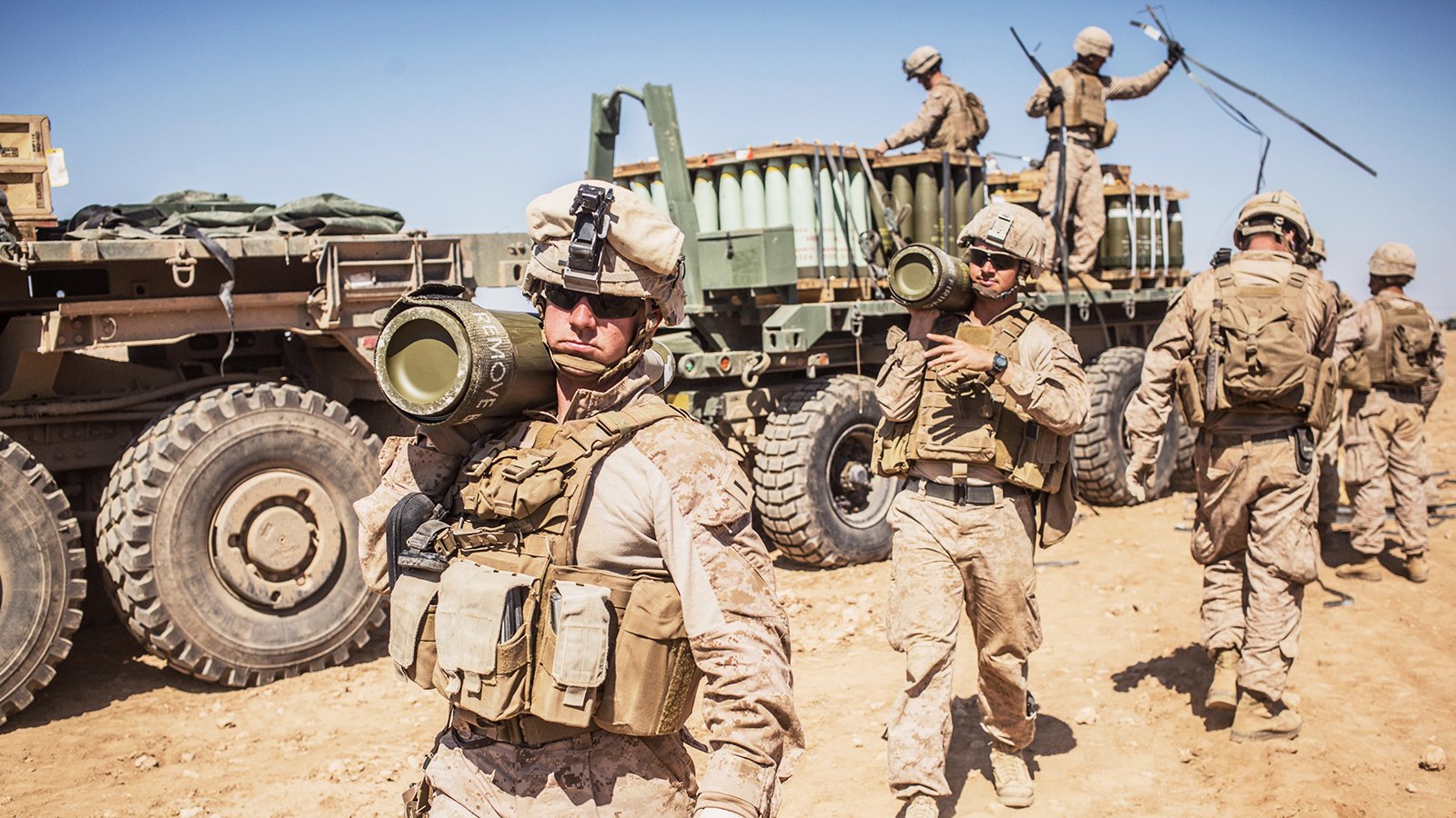 در این تصویر، گروهی از سربازان نظامی در حال فعالیت در یک محیط بیابانی دیده میشوند. آنها لباسهای نظامی به تن دارند و بخشی از تجهیزات سنگین را بارگیری میکنند. یکی از سربازان در پیشزمینه، موشک یا سلاحی را به دوش گرفته و به سمت دوربین نگاه میکند. در پسزمینه، وسیله نقلیه نظامی و دیگر سربازان مشغول به کار در حال حرکت هستند. آسمان آبی و صاف به همراه زمین خشک و شنزار نمایی از یک منطقه نظامی را به تصویر میکشد.