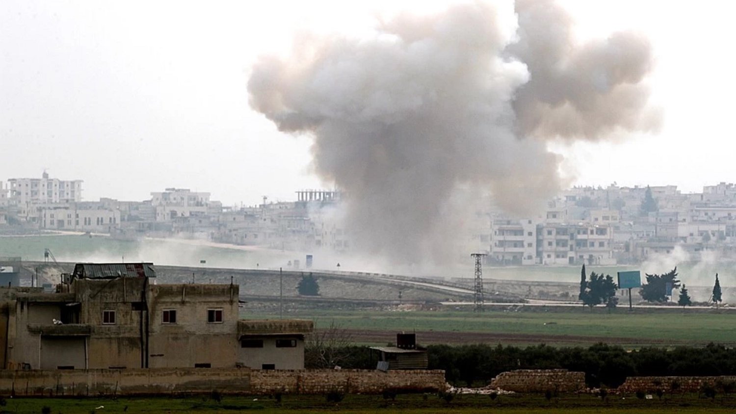 تصویر یک صحنه جنگی را نشان میدهد که در آن دود و گرد و غبار ناشی از یک انفجار دیده میشود. در پسزمینه، ساختمانهای شهری و یک منطقه باز مشاهده میشود. آسمان ابری است و حالت کلی تصویر به موقعیتی پر تنش و خطرناک اشاره دارد. دود در حال بلند شدن از زمین به سمت آسمان است و نشانههایی از ویرانی و نبرد در تصویر مشهود است.