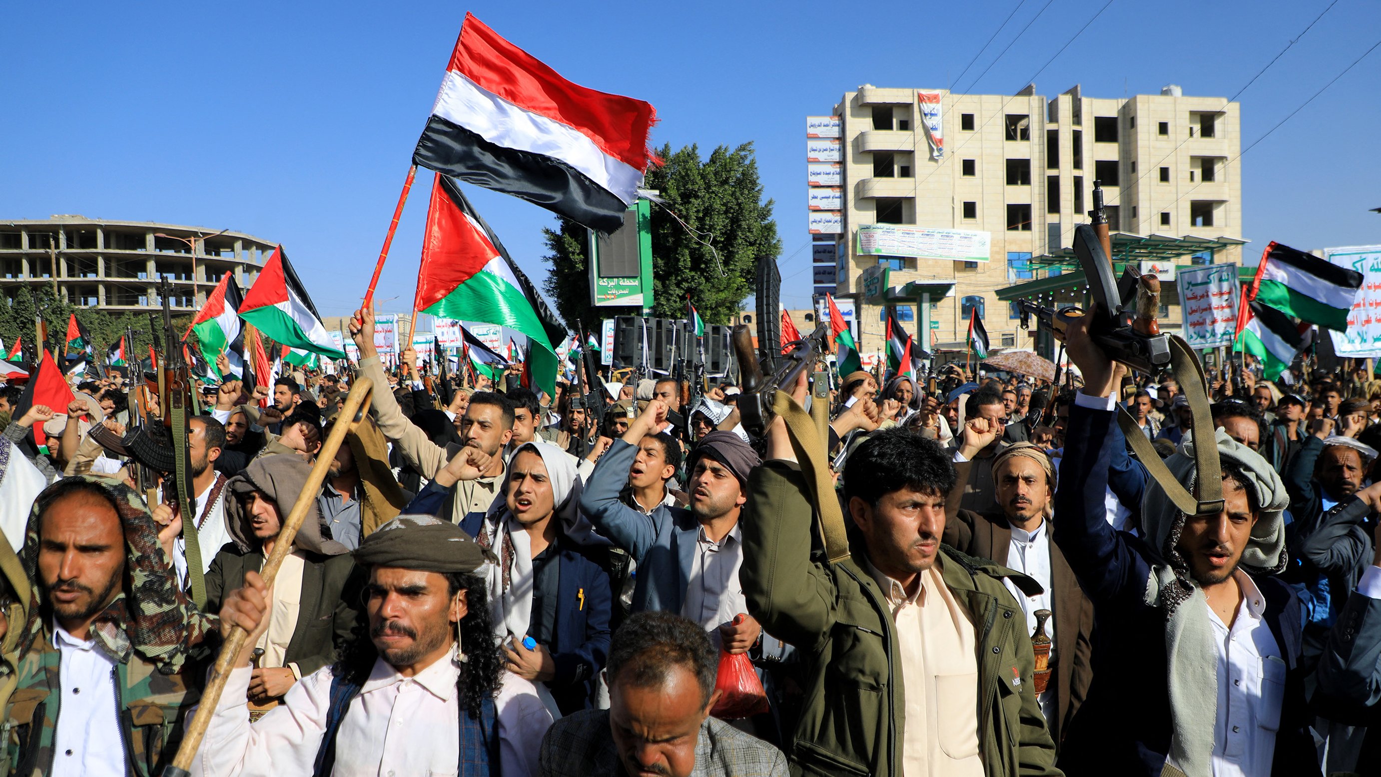 در این تصویر، تعداد زیادی از مردم در خیابانی تجمع کردهاند و پرچمهایی را در دست دارند. هر فردی ظاهری متعهد و با انرژی به نظر میرسد و برخی از آنها در حال فریاد زدن یا شعار دادن هستند. لباسها معمولاً سنتی هستند و برخی از شرکتکنندگان در حال بیان نظرات یا احساسات خود در مورد موضوعی خاص به نظر میرسند. محیط اطراف شامل ساختمانهای بلند و تابلوهای تبلیغاتی است که نشاندهنده یک میدان عمومی یا خیابان پرجنبوجوش است. پرچمها به رنگهای مختلف، خصوصاً رنگهای قرمز، سیاه و سفید، به خوبی دیده میشوند.