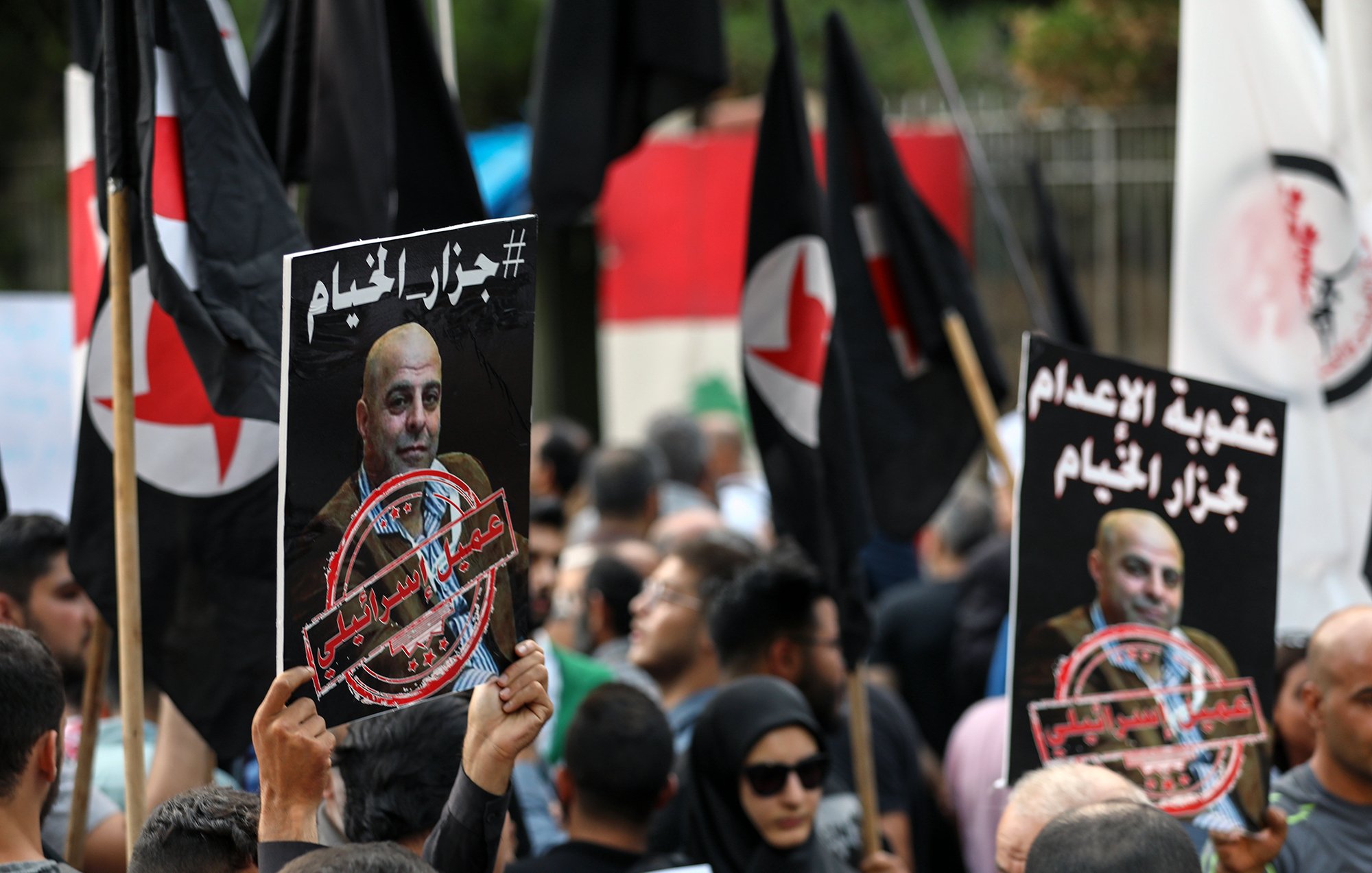 تصویر نشاندهنده یک تجمع اعتراضی است که مردم در آن پلاکاردهایی با نوشتههایی به زبان عربی و تصاویر شخصی را در دست دارند. در پسزمینه، پرچمهای سیاه رنگ با نمادهایی خاص به چشم میخورند. جمعیت به وضوح متنوع است و افراد مختلف در حال نمایش اعتراضات خود هستند. جو کلی تصویر به نظر معترضانه و پرشور میرسد.