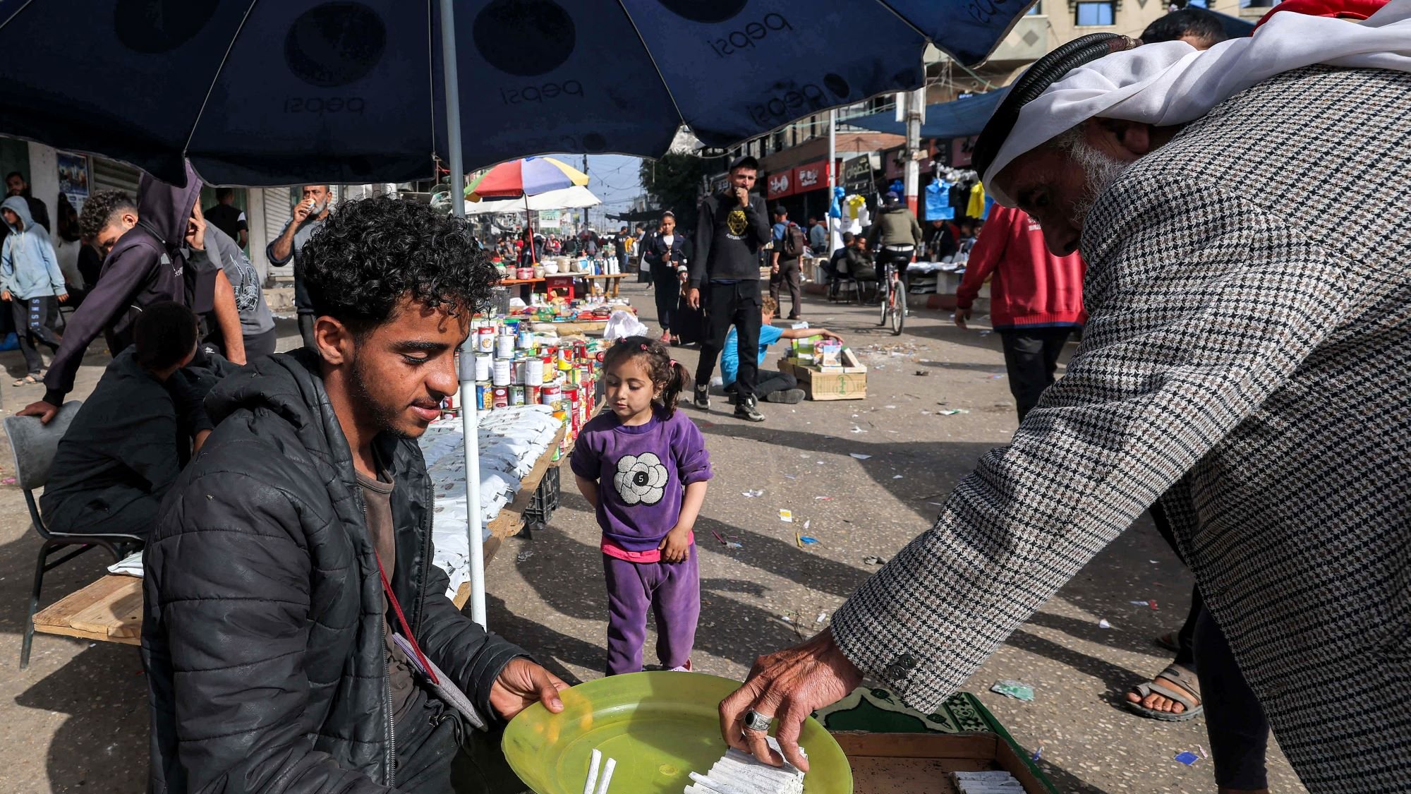 در تصویر، یک بازار محلی به چشم میخورد. در وسط تصویر، مرد جوانی نشسته و در حال فروش کالاهایی است. او یک بشقاب سبز رنگ را در دست دارد که در آن چند قلم وجود دارد. به نظر میرسد که او به مشتریاش توجه دارد که در حال انتخاب کالاهاست. در کنار آن، یک دختر بچه نیز در حال بازی کرده و به صحنه نگاه میکند. در پسزمینه، افراد دیگری در حال خرید و گشت و گذار هستند و جوی شلوغ و پرجنبوجوش به چشم میخورد. سایه چترهایی که بالای سر برخی افراد است، فضایی دلپذیر ایجاد کرده است.