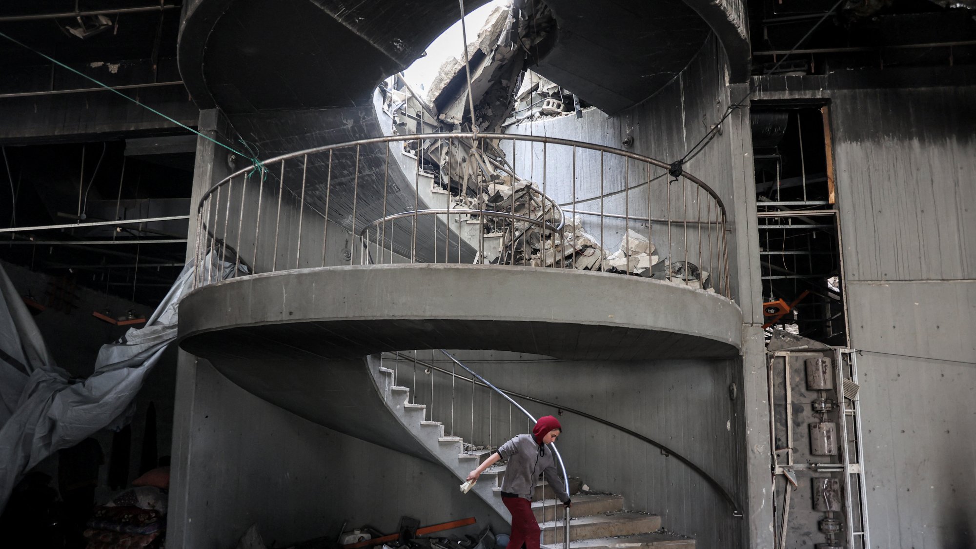 در این تصویر، یک پلهگردان دیده میشود که به دلیل تخریب، وضعیت نامناسبی دارد. بخشی از سقف بالای پلهها و دیوارها فرو ریخته و آوار زیادی در اطراف پلهها جمع شده است. یک فرد با عصا در حال حرکت در پلهها است و به نظر میرسد در تلاش است تا از این فضای خراب عبور کند. نور طبیعی از سقف خراب به داخل تابیده و فضایی دلگیر و غمانگیز به وجود آورده است.