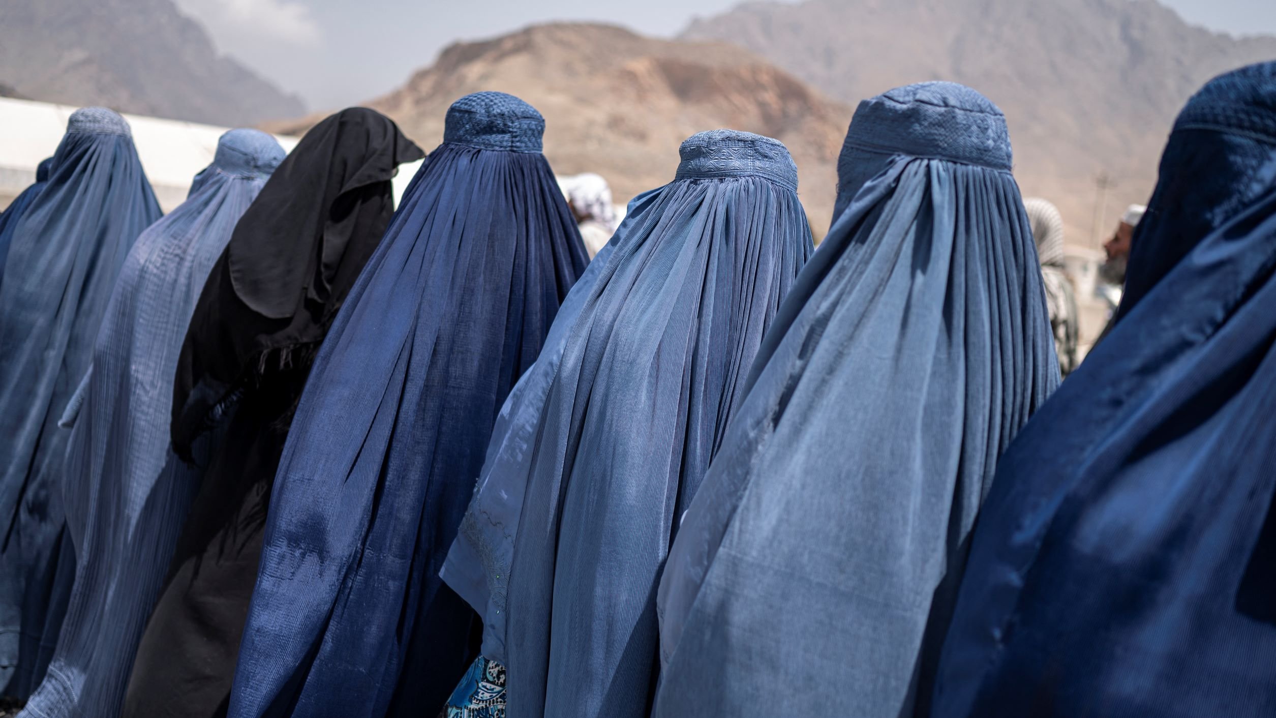 در این تصویر تعدادی زنان دیده میشوند که پوششهایی به رنگهای آبی و مشکی بر تن دارند. آنها از پشت به دوربین ایستادهاند و فقط قسمتهایی از پوششهایشان قابل مشاهده است. نمای背景 کوهها و فضای باز است، که ممکن است به منطقهای کوهستانی اشاره داشته باشد. این تصویر حس سنتی و فرهنگی خاصی را منتقل میکند.