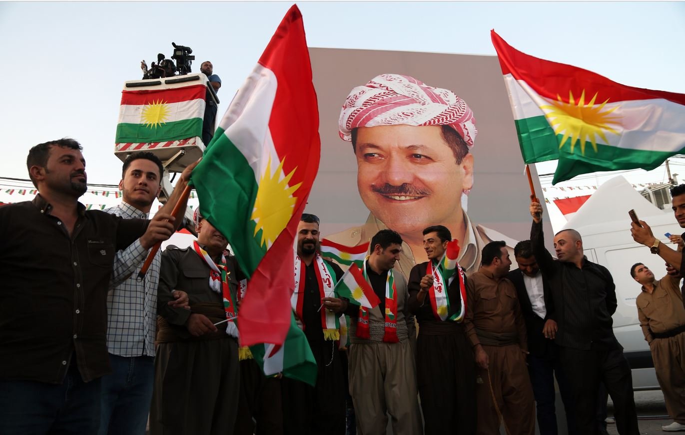 در تصویر گروهی از مردان لباس کردی به همراه پرچمهای کردستان را مشاهده میکنیم. آنها در حال ایستادن در مقابل یک بنر بزرگ هستند که تصویر فردی را نمایش میدهد. افراد با نشاط و خوشحالی پرچمها را در دست دارند و به نظر میرسد که در یک مراسم یا جشن خاص شرکت کردهاند. فضای جمعی و پر انرژی به خوبی در تصویر حس میشود.