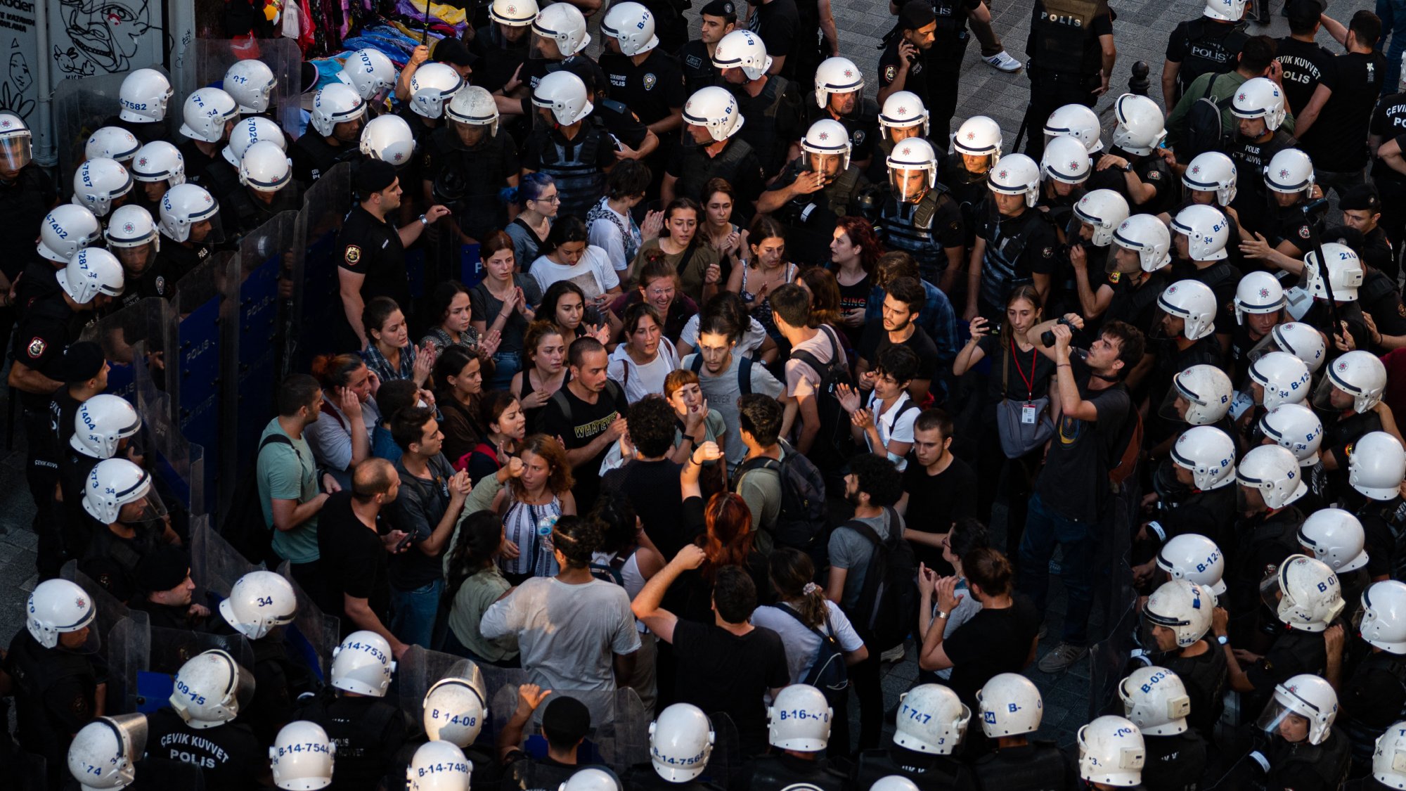 در این تصویر، گروهی از مردم در حال تظاهرات یا تجمعی هستند که با نیروهای پلیس محاصره شدهاند. افراد معترض در وسط تصویر قرار دارند و به نظر میرسد در حال گفتوگو یا گفتگو با پلیس هستند. نیروهای پلیس در اطراف آنها ایستادهاند و کلاههای مخصوصی به سر دارند. فضای تصویر نشاندهنده تنش و فعالیت اجتماعی است، با تجمع جمعیتی که به سمت نیروهای پلیس نگاه میکنند.