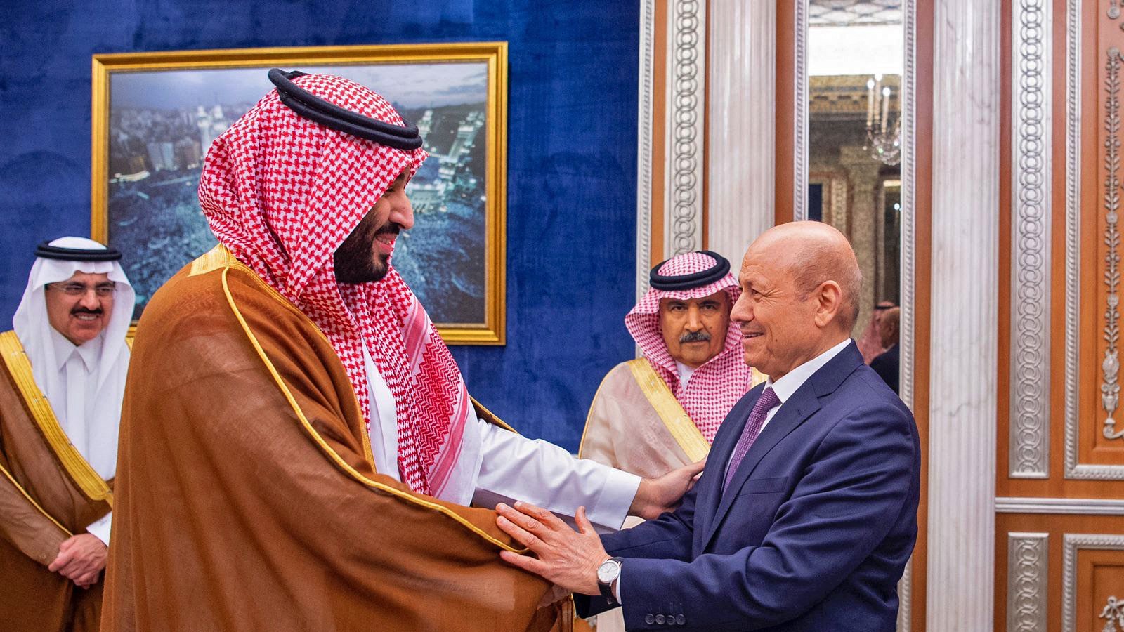 در این تصویر، دو مرد در حال گفتوگو و خوشآمدگویی به یکدیگر هستند. یکی از آنها لباس سنتی عربی به نام "ثوب" به تن دارد و چفیهای خاص بر سر دارد. او به نظر میرسد که در مقام رهبری قرار دارد. مرد دیگر نیز به صورت رسمی لباس پوشیده است و نشان از احترام و ادب در بیان حرکات او دیده میشود. در پسزمینه، دکوراسیون مجلل و لوکس با تزئینات طلایی و تابلوهایی از مناظر شهری دیده میشود که به فضای رسمی این ملاقات افزوده است.