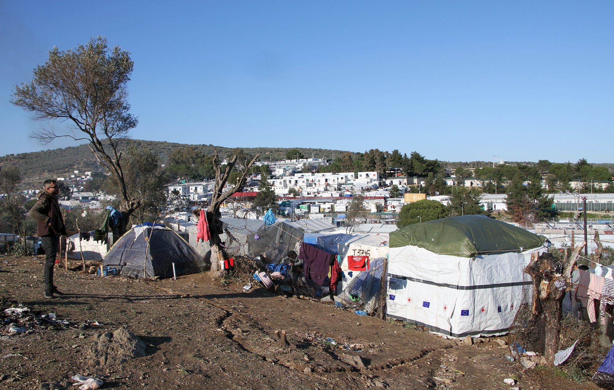 تصویر نشاندهنده یک منطقه با وضعیت نامناسب است که در آن چادرهایی به عنوان محل سکونت نصب شدهاند. در پسزمینه، زمینهای ناهموار و درختانی مشاهده میشود. پسزمینه شهری با ساختمانهایی دیده میشود که نسبتاً به چادرها نزدیک هستند. احساس کلی تصویر بیانگر شرایط سخت زندگی افرادی است که در این چادرها سکونت دارند. آسمان آبی و روز روشن است، اما وضعیت محیط نشاندهنده فقر و مشکلات معیشتی است.