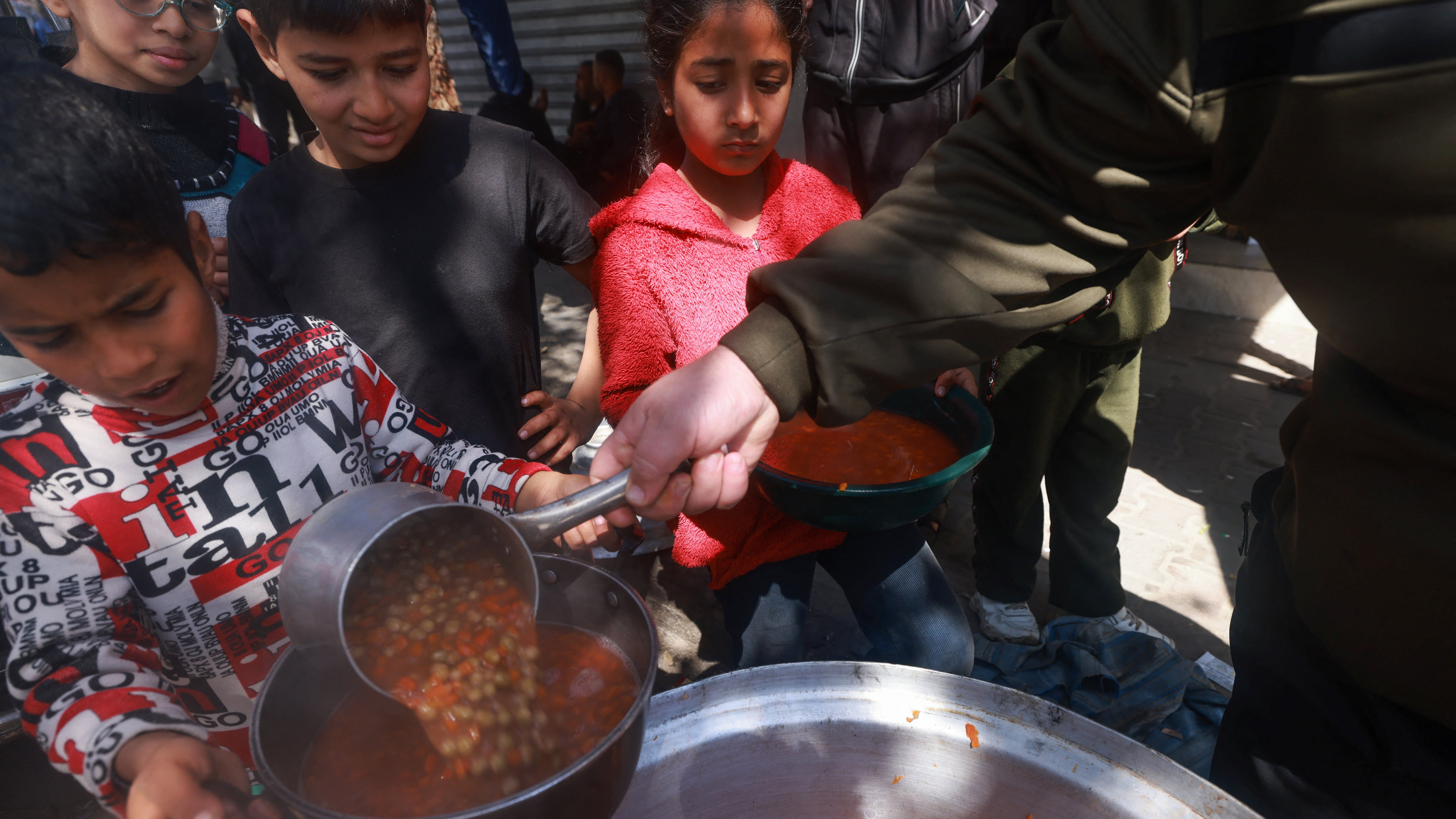 در این تصویر، گروهی از کودکان در حال دریافت غذا هستند. یکی از کودکان که لباسهای رنگارنگ دارد، با دقت در حال ریختن خورش یا سوپ به ظرفش است. در پسزمینه، سایر کودکان نیز منتظر هستند تا نوبتشان برسد. محیط بیرونی به نظر شلوغ و غیررسمی است و نشاندهنده یک فعالیت جمعی برای سرو غذا به افراد نیازمند میباشد.