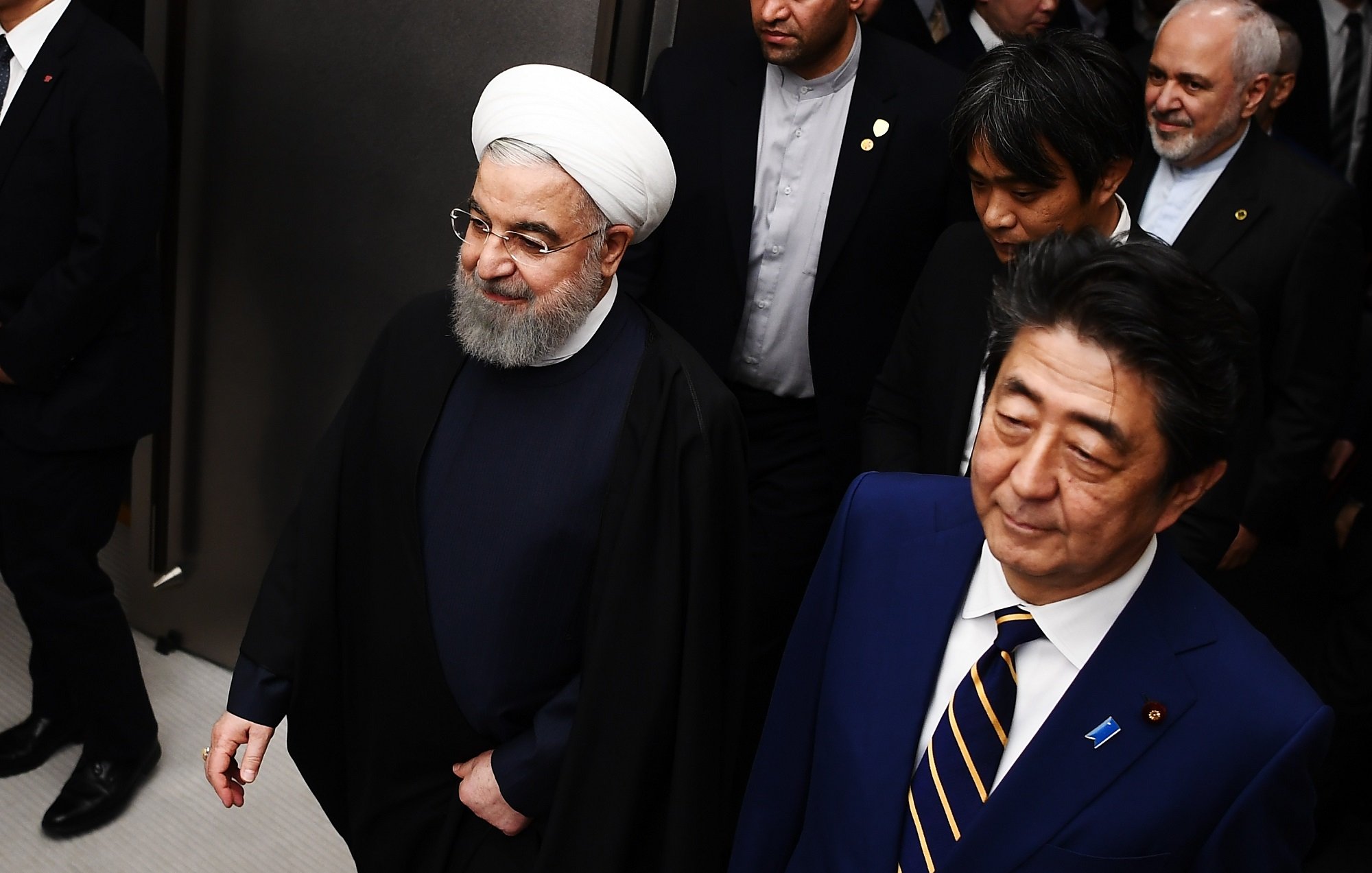 در تصویر، دو مرد در حال پیادهروی هستند. یکی از آنها دارای پوشش سنتی ایرانی با عمامه سفید و جبه است، و به نظر میرسد که او مقام رسمی و بلندپایهای باشد. فرد دیگر در کنار او، با کت و شلوار رسمی و کراوات، لبخند به لب دارد. در پسزمینه، چند نفر دیگر نیز دیده میشوند که به نظر میرسد همراهان آنها هستند. محیط تصویر ممکن است یک کنفرانس یا جلسه رسمی را نشان دهد.