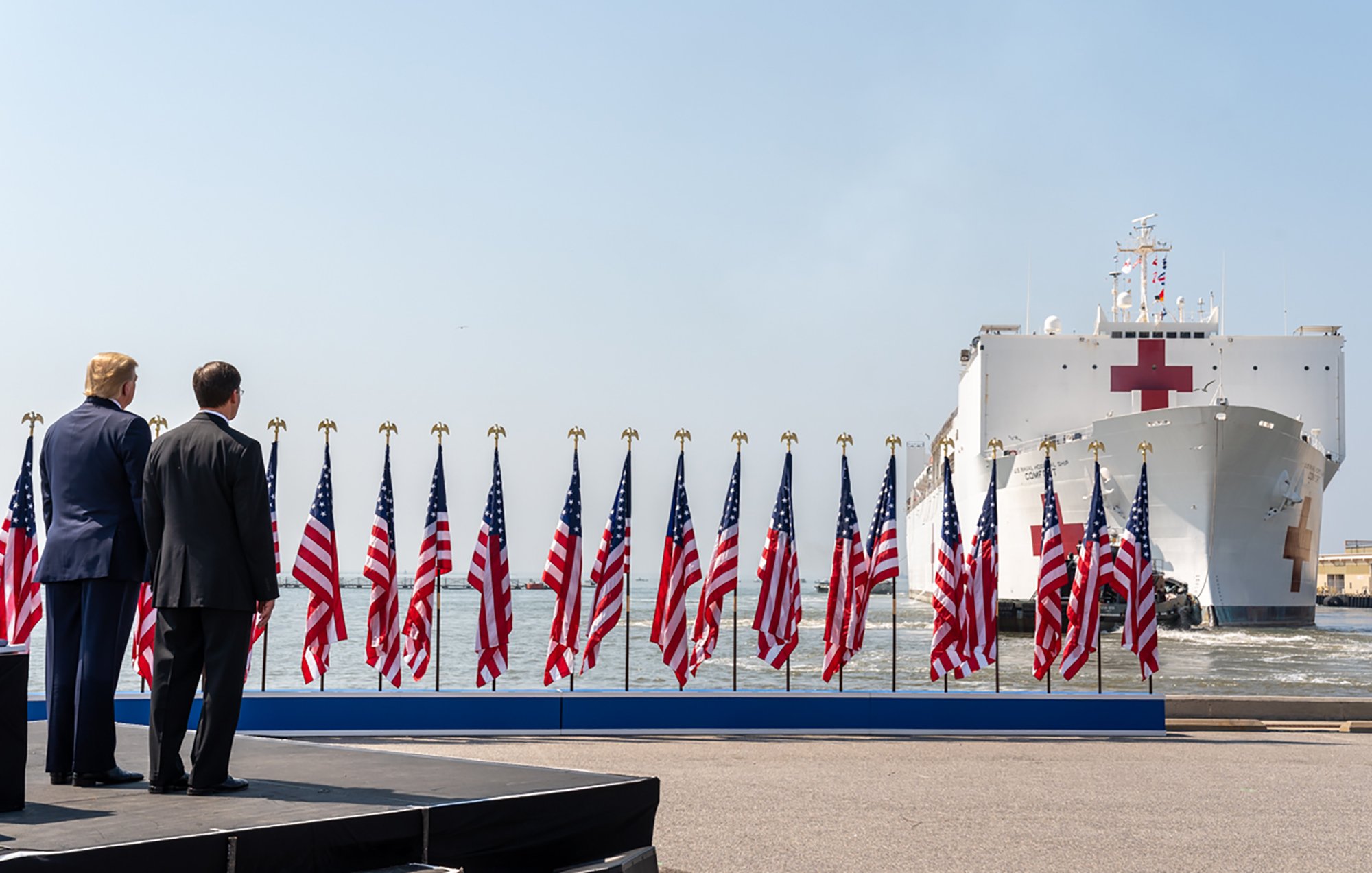 در این تصویر، دو مرد در حال ایستادن هستند که به یک کشتی بزرگ با علامت صلیب قرمز نگاه میکنند. کشتی در حال ورود به بندر است و در اطراف آن چندین پرچم ایالات متحده در صف ایستادهاند. آسمان آبی و واضح است و فضای عمومی شلوغ به نظر میرسد. مردان در نزدیکی یک صحنه یا سکو قرار دارند که احتمالاً برای یک رویداد یا مراسم خاص آماده شده است.