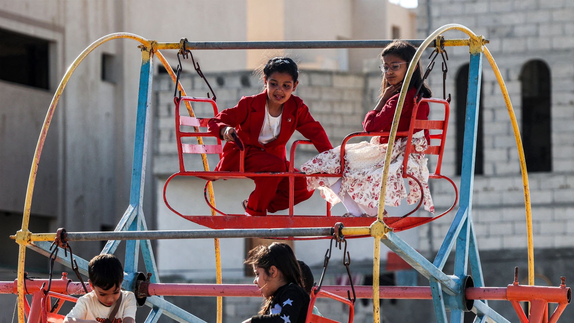 در تصویر، چند کودک در یک وسیله بازی در پارک دیده میشوند. وسیله بازی شبیه یک چرخوفلک است و رنگهای روشنی دارد. دو کودک در بالای چرخوفلک نشستهاند؛ یک پسر با لباس قرمز و یک دختر با لباس سفید و گلدار. در پایین نیز تعدادی کودک دیگر در حال بازی و نشستن هستند. فضای تصویر دارای حس شادی و نشاط است و نشاندهنده لحظات خوشی از دوران کودکی است. زمینه تصویر ساختمانهایی با نمای ساده به چشم میخورد.