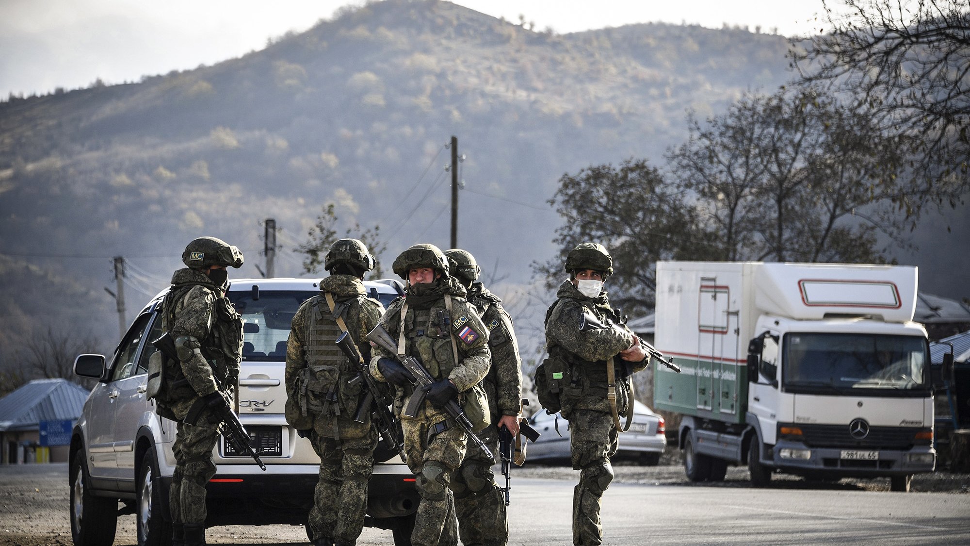 تصویر گروهی از سربازان مسلح را نشان میدهد که در یک منطقه روستایی قرار دارند. سربازان در لباسهای نظامی و کلاههای ایمنی به وضوح مسلح هستند و در حال برقراری امنیت به نظر میرسند. پسزمینه تصویر شامل کوهها و درختان است که به فضا حالت طبیعی میدهد. همچنین، چندین وسیله نقلیه در کنار جاده دیده میشوند. نور تصویر نیز نشاندهنده روز روشن است.