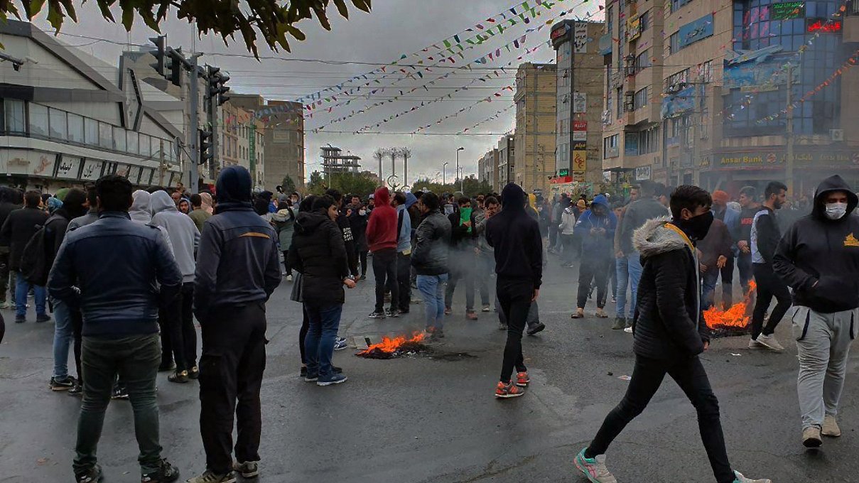 تصویر نشاندهنده یک تجمع اعتراضی در یک خیابان است. در پسزمینه، تعدادی از ساختمانها دیده میشود و آسمان به رنگ خاکستری است. جمعیت زیادی از افراد در لباسهای گرم در حال ایستادن یا حرکت کردن هستند. برخی از آنها ماسک به چهره دارند. در چند نقطه از خیابان، دود و شعلههایی به چشم میخورد که نشاندهنده وجود آتش است. همچنین، تزئینات و پرچمهایی در بالای خیابان دیده میشود که به نظر میرسد نشاندهنده یک مناسبت خاص یا جشنواره است. جمعیت به نظر میرسد که در حال تظاهرات یا اعتراض هستند.