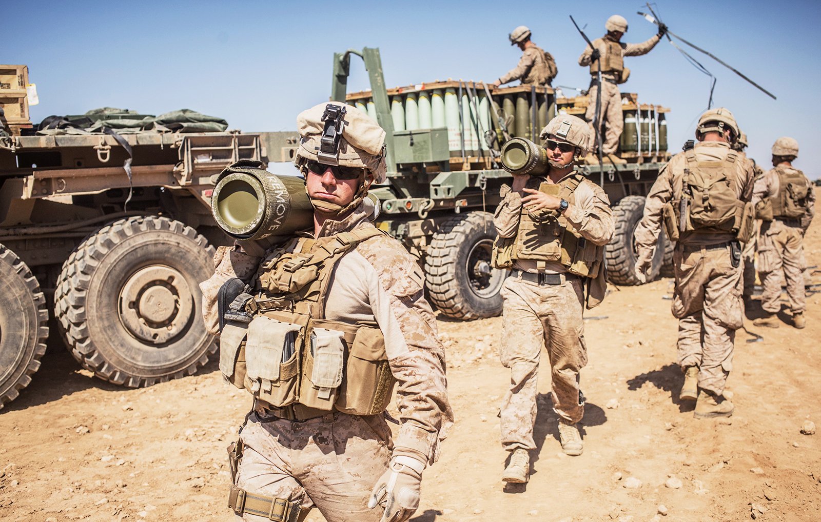 در این تصویر، گروهی از سربازان نظامی در حال فعالیت در یک محیط بیابانی دیده میشوند. آنها لباسهای نظامی به تن دارند و بخشی از تجهیزات سنگین را بارگیری میکنند. یکی از سربازان در پیشزمینه، موشک یا سلاحی را به دوش گرفته و به سمت دوربین نگاه میکند. در پسزمینه، وسیله نقلیه نظامی و دیگر سربازان مشغول به کار در حال حرکت هستند. آسمان آبی و صاف به همراه زمین خشک و شنزار نمایی از یک منطقه نظامی را به تصویر میکشد.