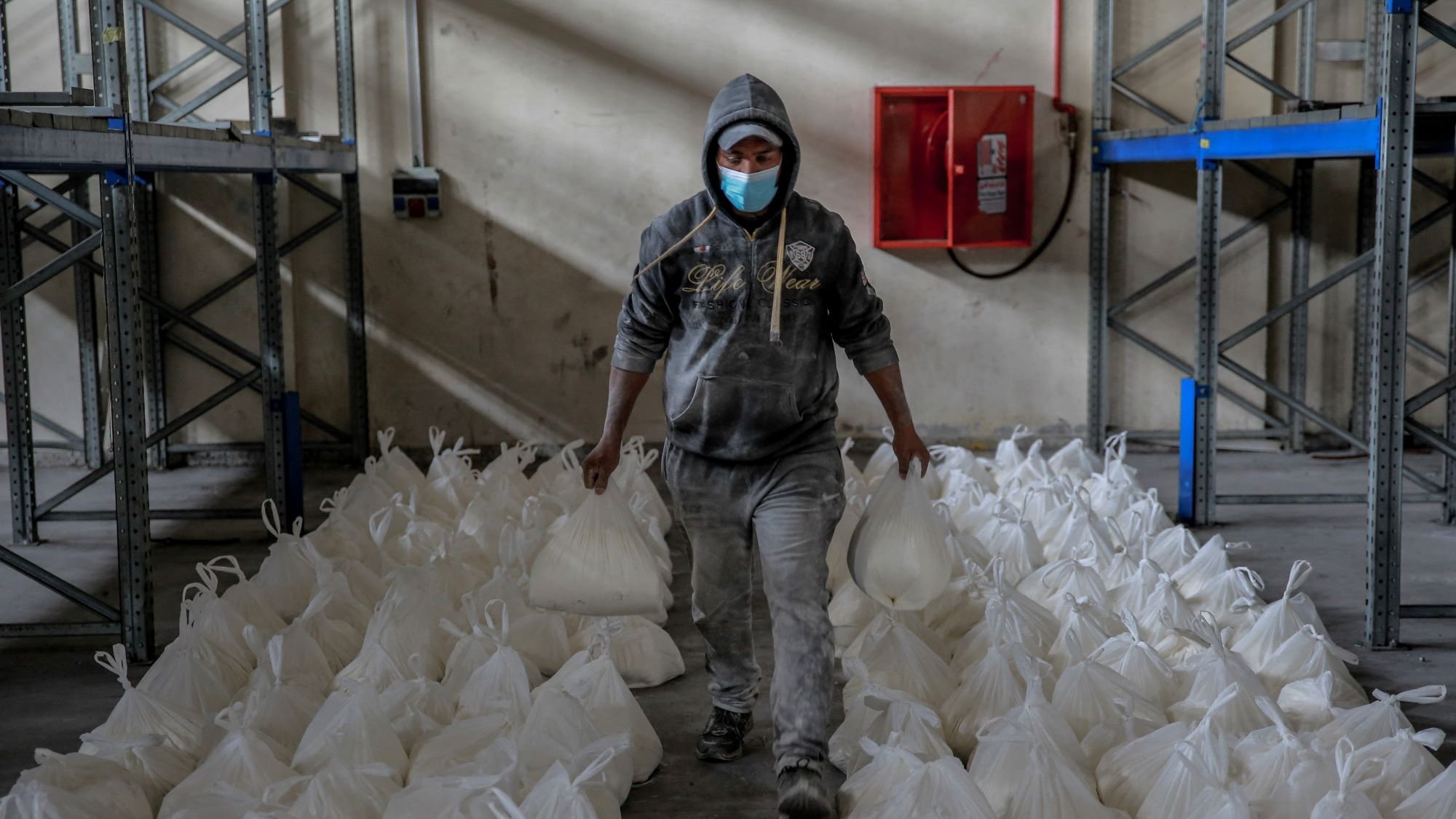 در این تصویر، یک مرد با پوشش خاکستری و کلاهی بر سر، در حال حرکت در یک محل انبار است. او یک ماسک به صورت دارد و در دو دست خود کیسههای سفیدی را حمل میکند. اطراف او ردیفهایی از کیسههای مشابه به طور منظم روی زمین قرار دارند. دیوارها و قفسههای فلزی در پسزمینه قابل مشاهده هستند که به نظر میرسد برای نگهداری کالاها استفاده میشوند. نور کمی در فضا وجود دارد و جو عمومی، فعالیتی را در زمینه توزیع یا انبارداری نشان میدهد.
