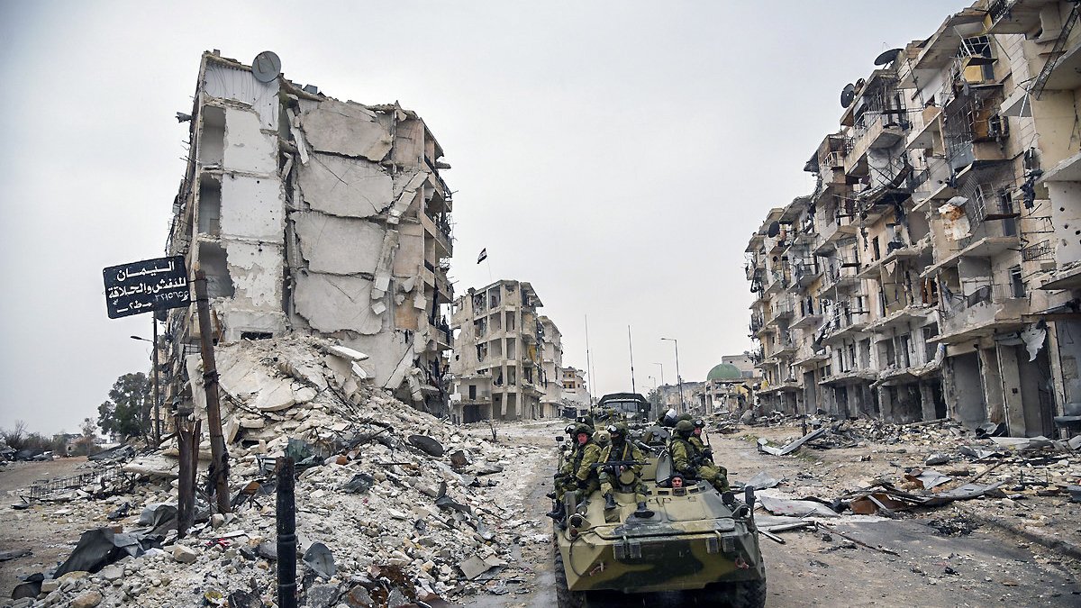 تصویر نشاندهندهٔ یک منطقهٔ ویران شده است که در آن چند ساختمان تخریب شده و در حال فرو ریختن به نظر میرسند. در این صحنه، یک وسیله نقلیه نظامی در حال حرکت است و تعدادی سرباز در آن نشستهاند. بقیهٔ محیط شامل زبالهها و نشانههای ویرانی است که به وضعیت بحرانی منطقه اشاره دارد. هوا ابری و تیره به نظر میرسد و فضایی خالی و ساکت را ایجاد کرده است.