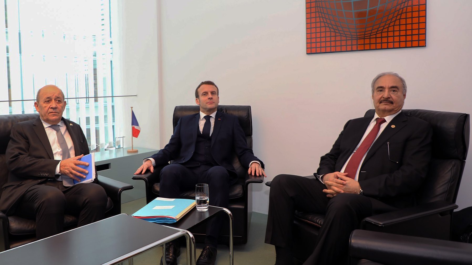 در این تصویر سه نفر نشستهاند. یکی از آنها در سمت چپ یک مرد با موهای خاکستری و در حال صحبت است. در وسط، مردی با کت و کراوات تیره نشسته که به نظر میرسد به موضوعی توجه میکند. شخص سوم در سمت راست یک مرد با موهای سیاه و چهرهای جدی است که او نیز در حال حاضر در نشستی قرار دارد. پسزمینه تصویر شامل پنجرههایی است که نور طبیعی را به اتاق وارد میکند. در کنار این افراد، پرچمی از فرانسه به چشم میخورد.