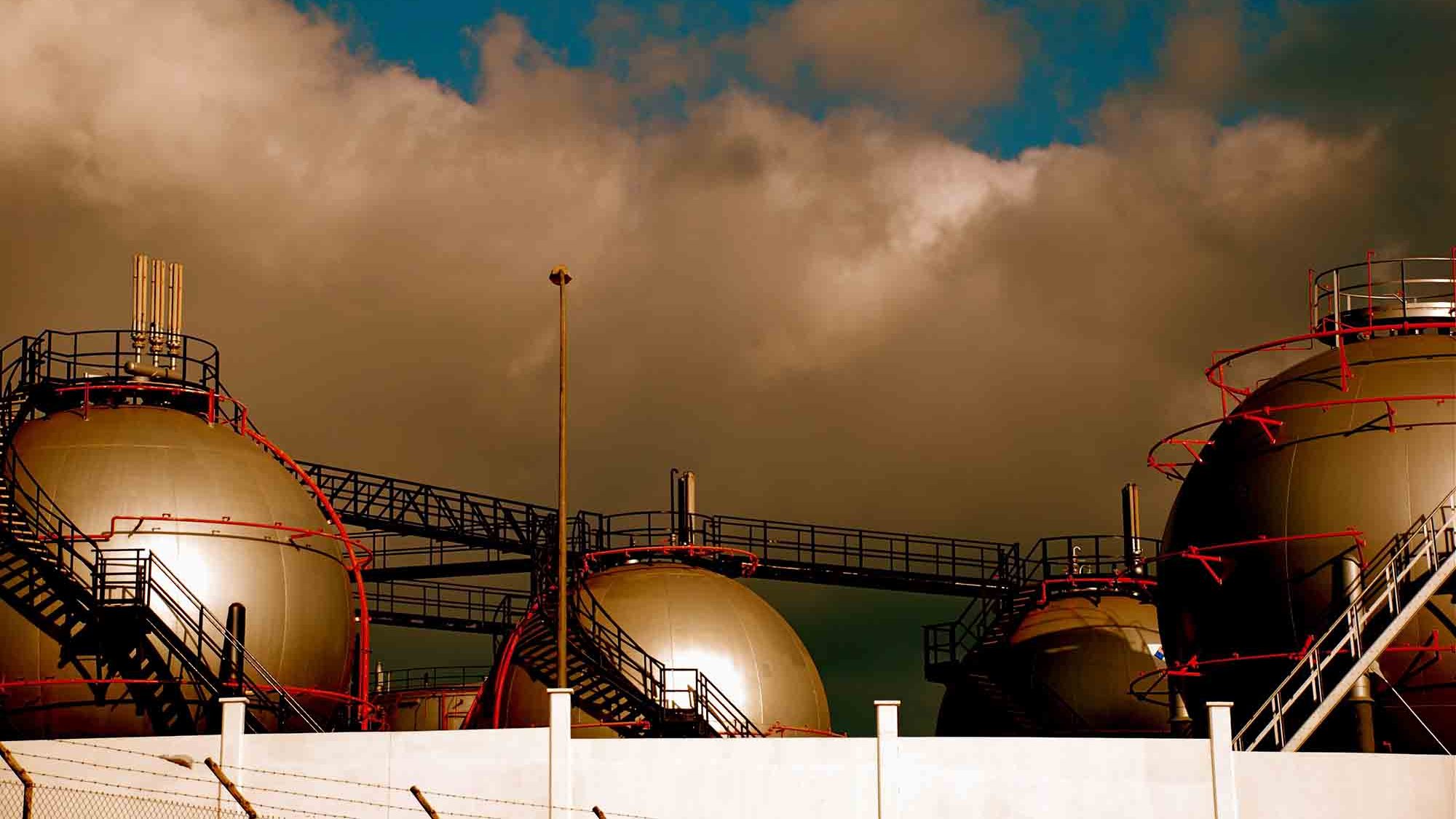 این تصویر نمایی از یک تأسیسات صنعتی را نشان میدهد که شامل چند مخزن بزرگ به شکل کروی است. مخازن روی سکویی قرار دارند و برخی از آنها داکتها و لولههای پیچیدهای دارند که به آنها متصل شدهاند. آسمان در پسزمینه کمی ابری است و رنگها به طور کلی گرم و متنوع به نظر میرسند. اجزای صنعت و بافتهای مختلف تأسیسات به خوبی در تصویر دیده میشوند.