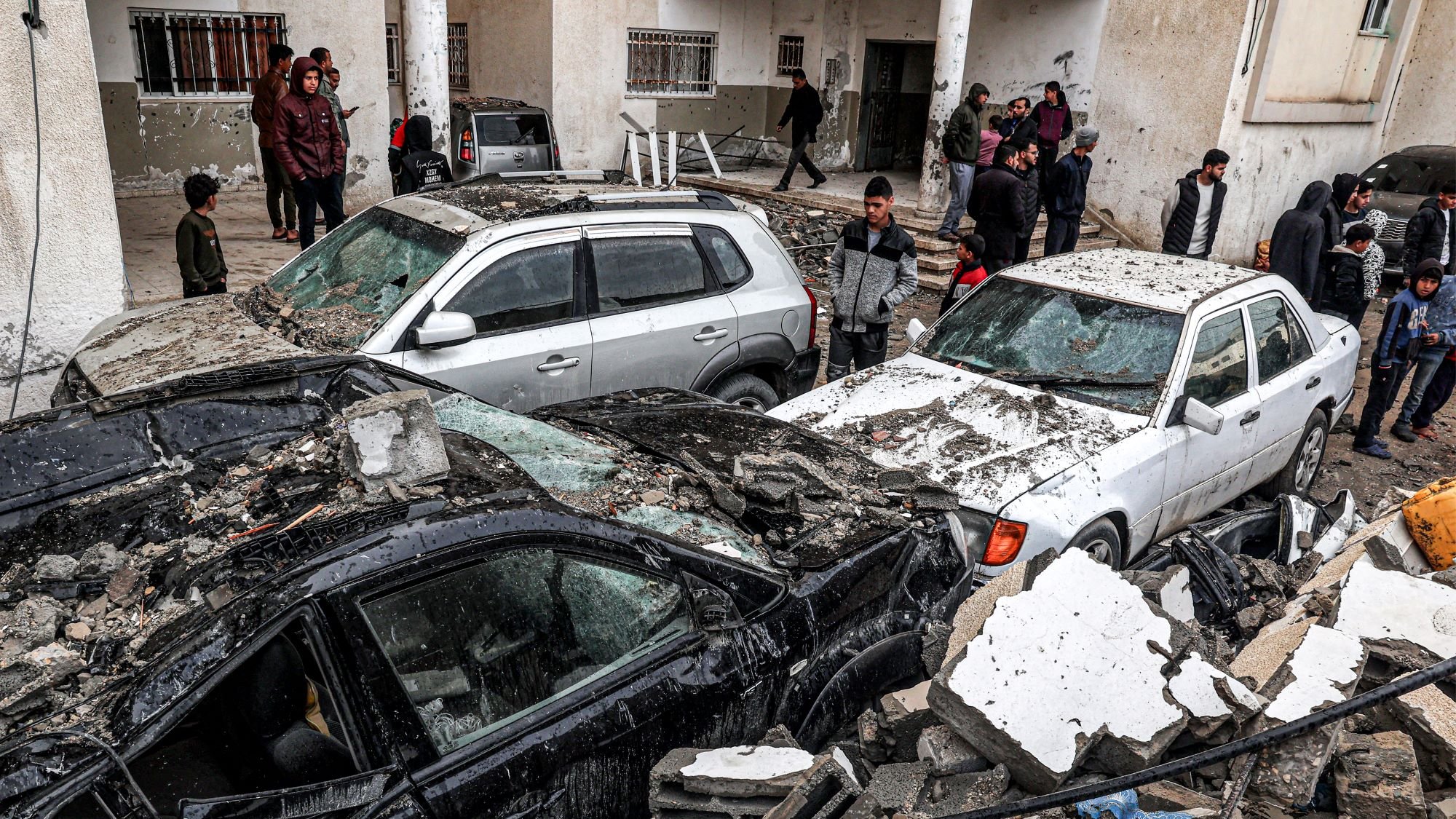 این تصویر نشاندهنده یک صحنه ویران شده به واسطه زلزله است. چندین خودرو زیر آوارهای ساختمانی قرار گرفتهاند و آسیب زیادی دیدهاند. افرادی در پسزمینه در حال مشاهده و بررسی وضعیت هستند. آوار خانهها و دیوارهای فروریخته در اطراف خودروها به چشم میخورد. به طور کلی، جو تصویر نشاندهنده اضطراب و فاجعه ناشی از زلزله است.