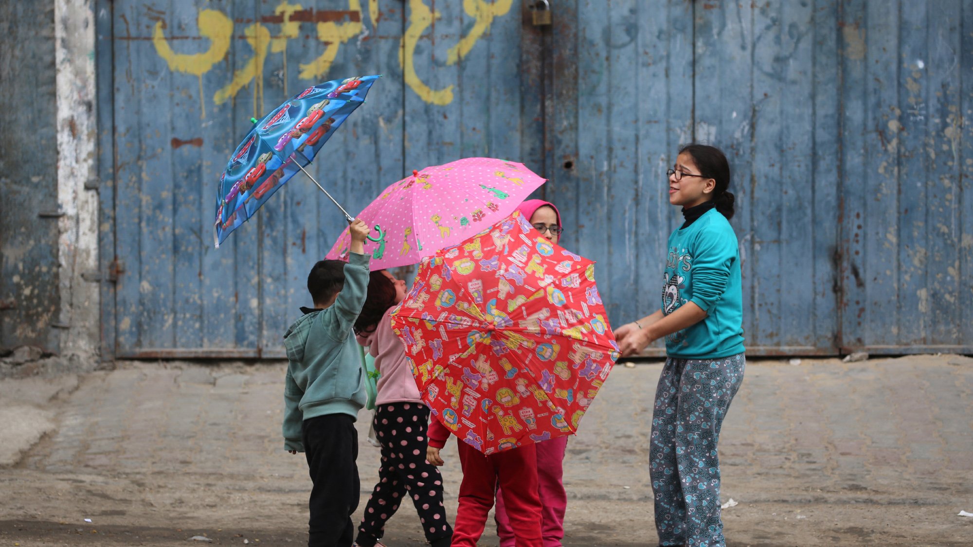در تصویر، چند کودک در حال بازی با چترهای رنگارنگ هستند. چترها طرحهای شاد و جذابی دارند و کودکان در حالی که در دایرهای ایستادهاند، به هم نزدیک هستند. زمینه تصویر یک دیوار آبی رنگ و زمین سنگفرش شده است.