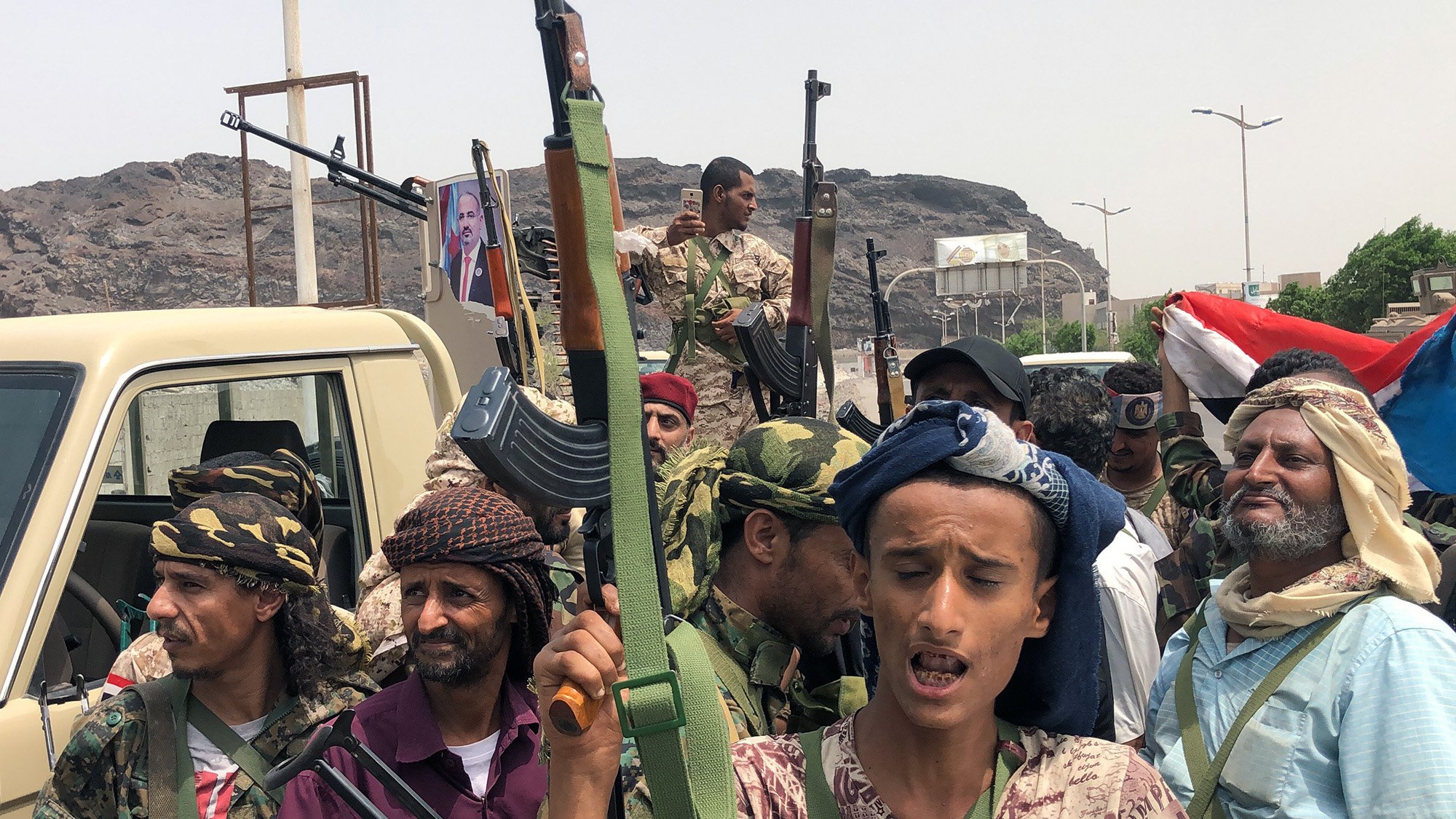 در این تصویر گروهی از مردان مسلح در یک منطقه عمومی دیده میشوند. برخی از آنها کلاهی به سر دارند و در دست سلاحهایی نظیر کلاشنیکف نگه داشتهاند. در پسزمینه، تصاویر و پرچمهایی از یمن قابل مشاهده است. به نظر میرسد آنها در حال برگزاری یک گردهمایی یا نمایش نیرو هستند. زمینه کوهستانی و ساختمانی در دوردست قرار دارد. جایگاههای زیادی از سوی خودروها در اطراف آنها وجود دارد.