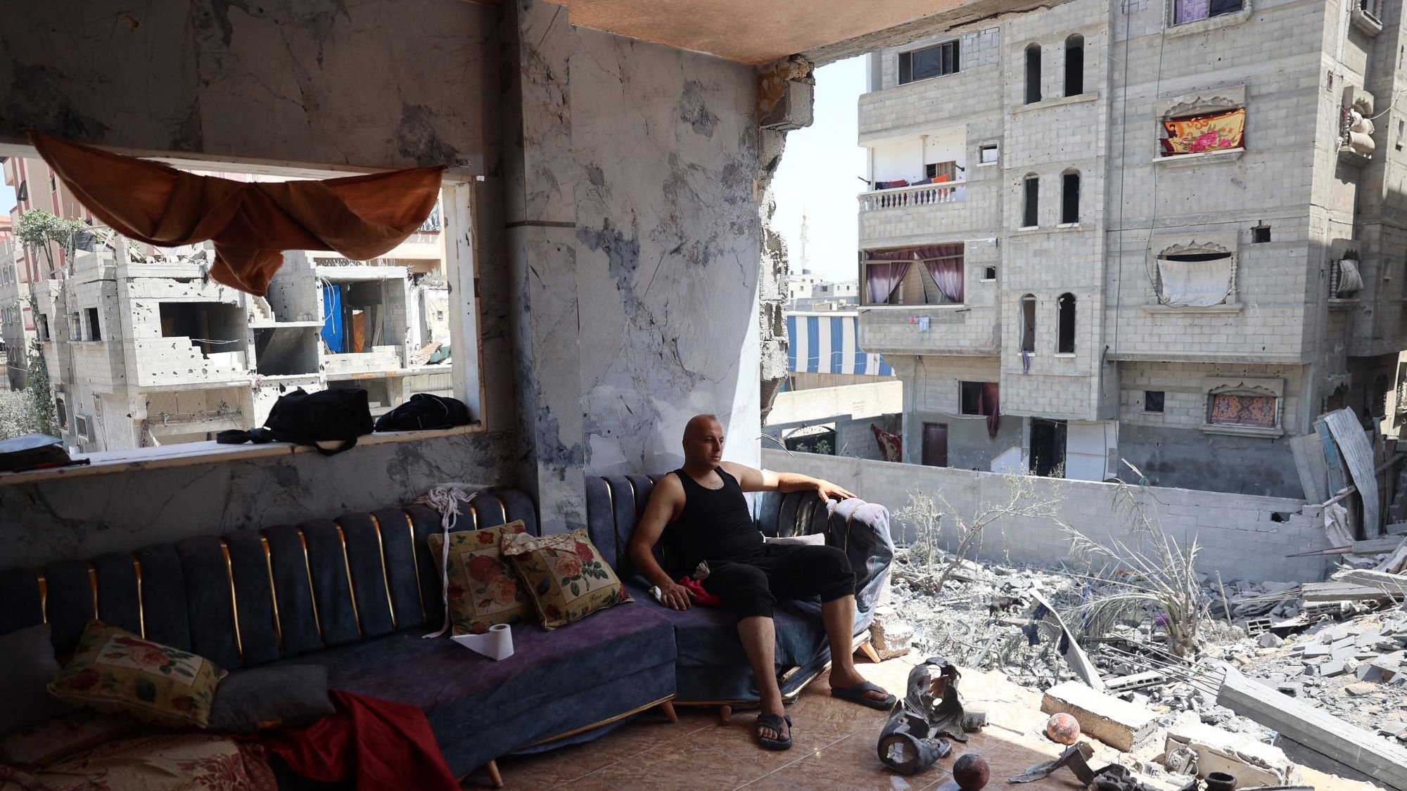 این تصویر یک فضا را نشان میدهد که دچار تخریب شدید شده است. در سمت راست، ساختمانهایی با دیوارهای آسیبدیده و پنجرههای شکسته دیده میشود. در سمت چپ، یک مرد در حال نشستن روی مبل درون یک اتاق است. او لباس سادهای به تن دارد و در کنار او وسایلی مانند کوسنها و اشیاء دیگر دیده میشود. فضای اتاق نیز به نظر میرسد که نشاندهنده شرایط نابسامان زندگی در آن منطقه باشد، با دیوارهایی که شکافها و آسیبهای واضحی دارند.整体اً، تصویر حس ناامیدی و خطر را منتقل میکند.