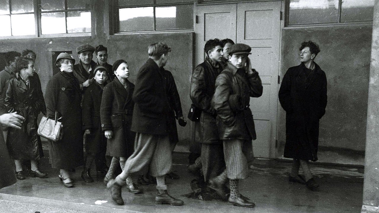 این تصویر سیاهوسفید گروهی از افراد را نشان میدهد که در یک محیط داخلی در حال حرکت هستند. برخی از افراد لباسهای گرم و کلوفت به تن دارند و به نظر میرسد که در حال عبور از یک فضای بسته هستند. تعدادی از افراد در حال گفتگو یا مراقب هستند، در حالی که دیگران به جلو نگاه میکنند. فضای تصویر حالت جدی و متفکری دارد و به طور کلی جو نگرانکنندهای را منتقل میکند.